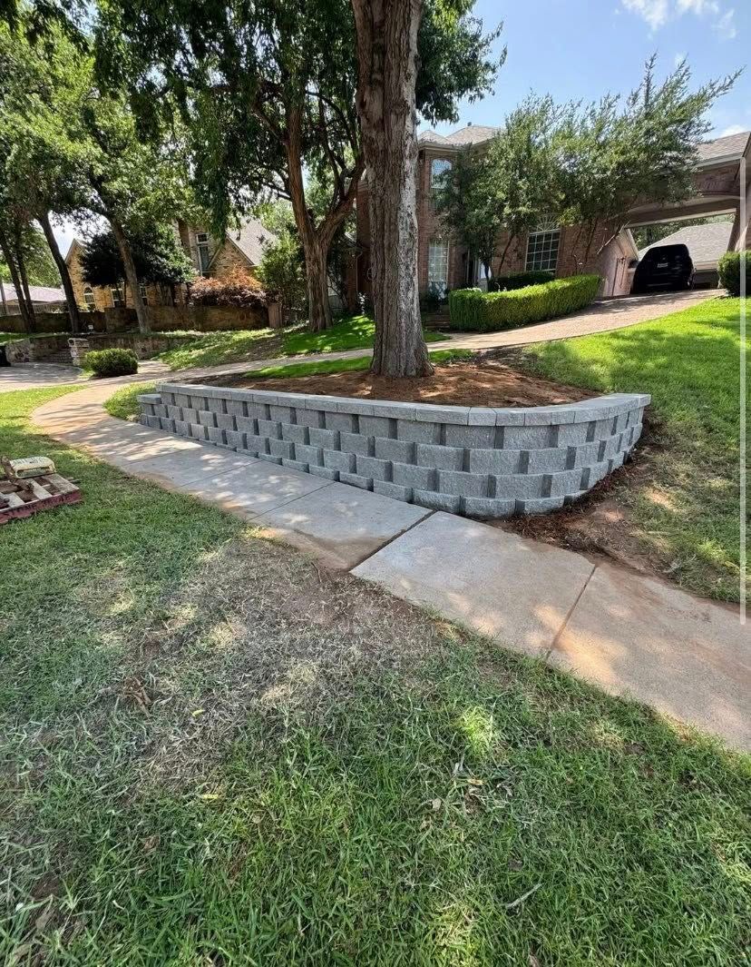 A retaining wall made of gray blocks surrounds a tree on a sidewalk near a house.
