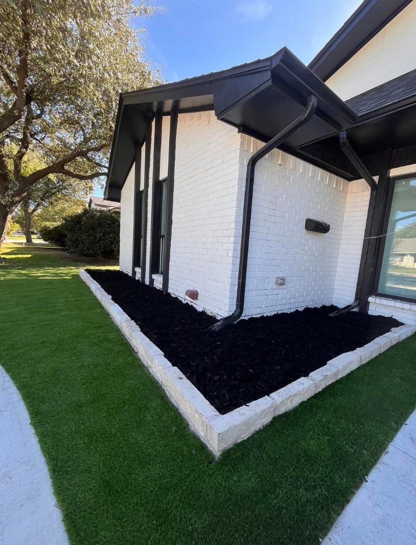 Corner of a house with white brick, black trim, and black mulch in a stone-bordered bed on green grass.