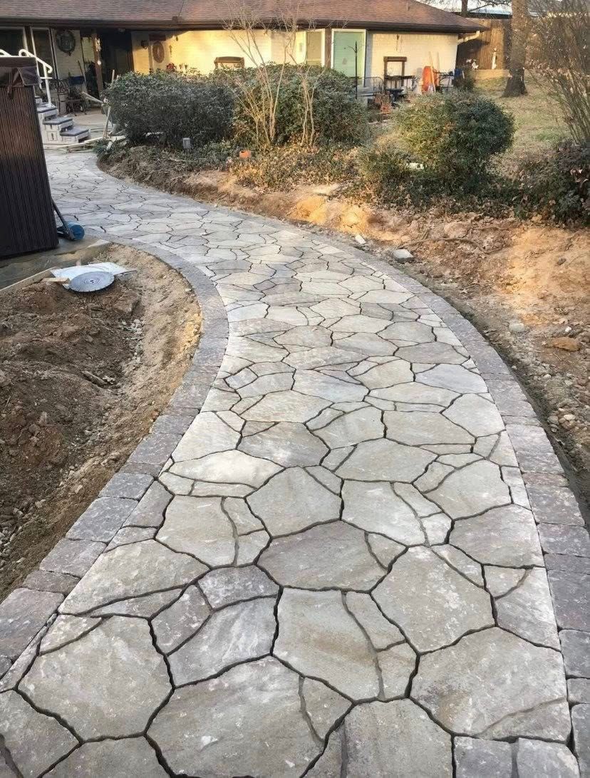 Stone walkway with a brick border curves towards a house, with landscaping on both sides.
