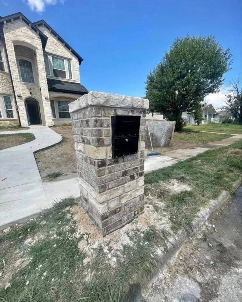 Stone mailbox post with black mail slot, beside a sidewalk and house.