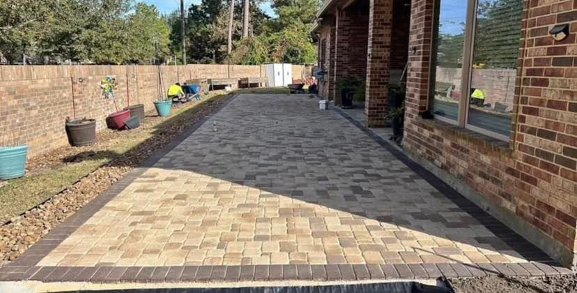 Brick patio with border adjacent to a brick house and a fenced yard with landscaping.