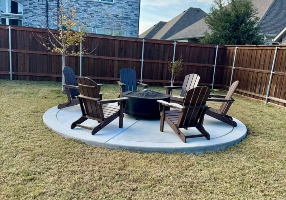 Fire pit with Adirondack chairs on a concrete patio in a backyard with a wooden fence.