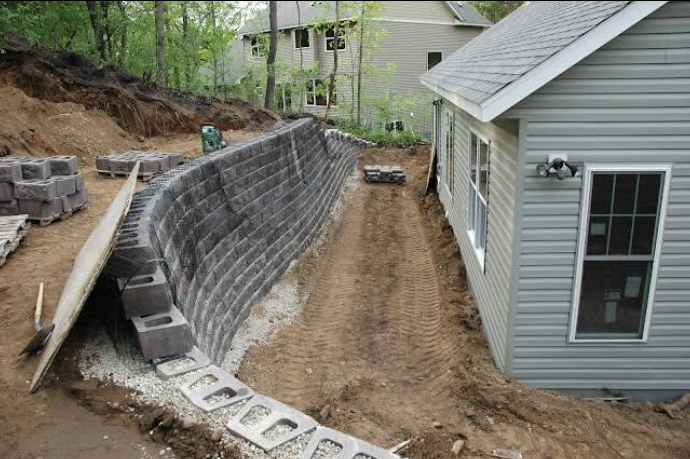 A retaining wall being built beside a house. Gray blocks and dirt on the ground.