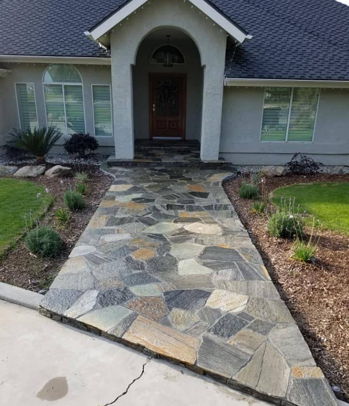 Stone pathway leading to a house with a brown door and gray siding, flanked by greenery.