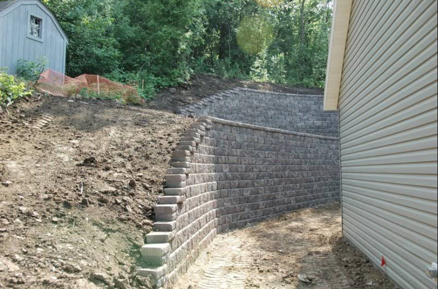 Stone retaining wall with steps on a hillside next to a house. Dirt and a small shed in the background.