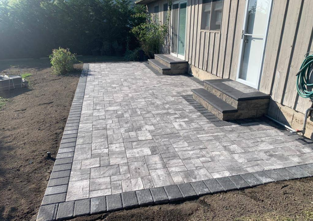 Paver patio with gray bricks and dark border, adjacent to a house with steps.