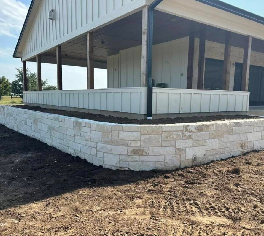 Stone retaining wall supporting a white farmhouse porch. Earth and blue sky in the background.