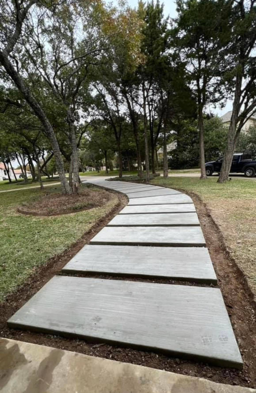 Concrete walkway with large rectangular pavers winding through a grassy area and trees.