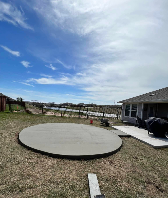 Circular concrete patio in a backyard under a blue sky, next to an existing patio.