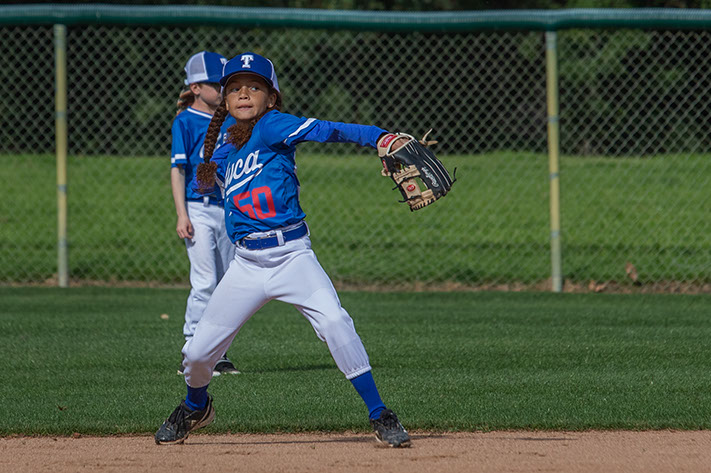 Girl in blue baseball uniform throwing a ball on a green field.