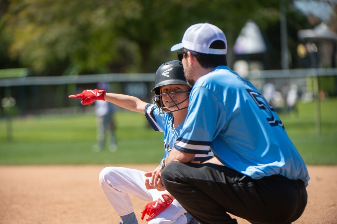 Baseball coach pointing; player in helmet on a base; both wearing light blue uniforms on a sunny field.