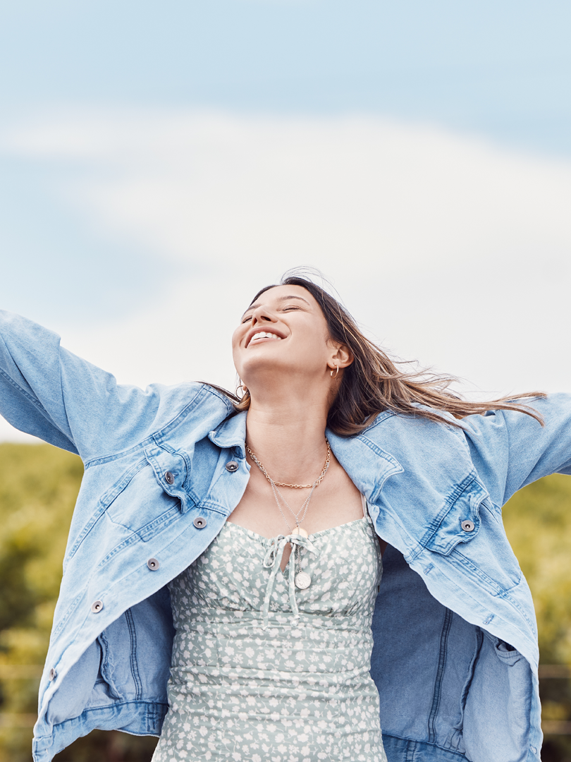 A woman in a dress and denim jacket is standing in a field with her arms outstretched.