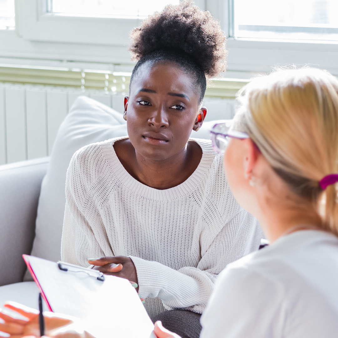 A woman is sitting on a couch talking to another woman while holding a clipboard.