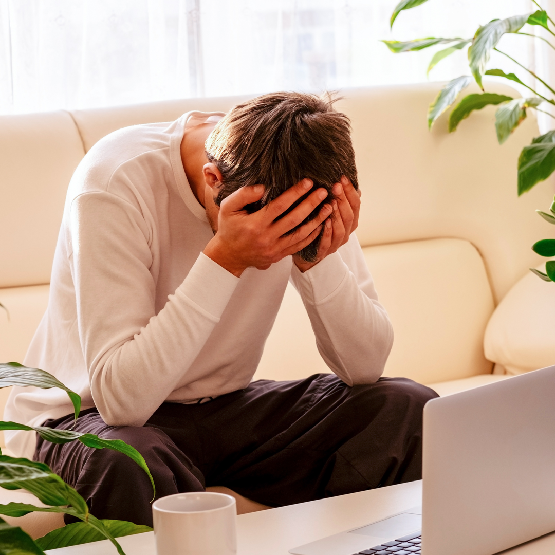 A man is sitting on a couch with his head in his hands in front of a laptop