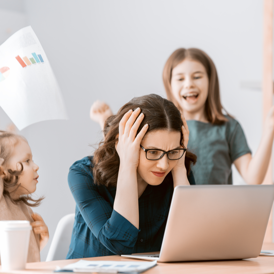 A woman is sitting at a table with a laptop and two children behind her.