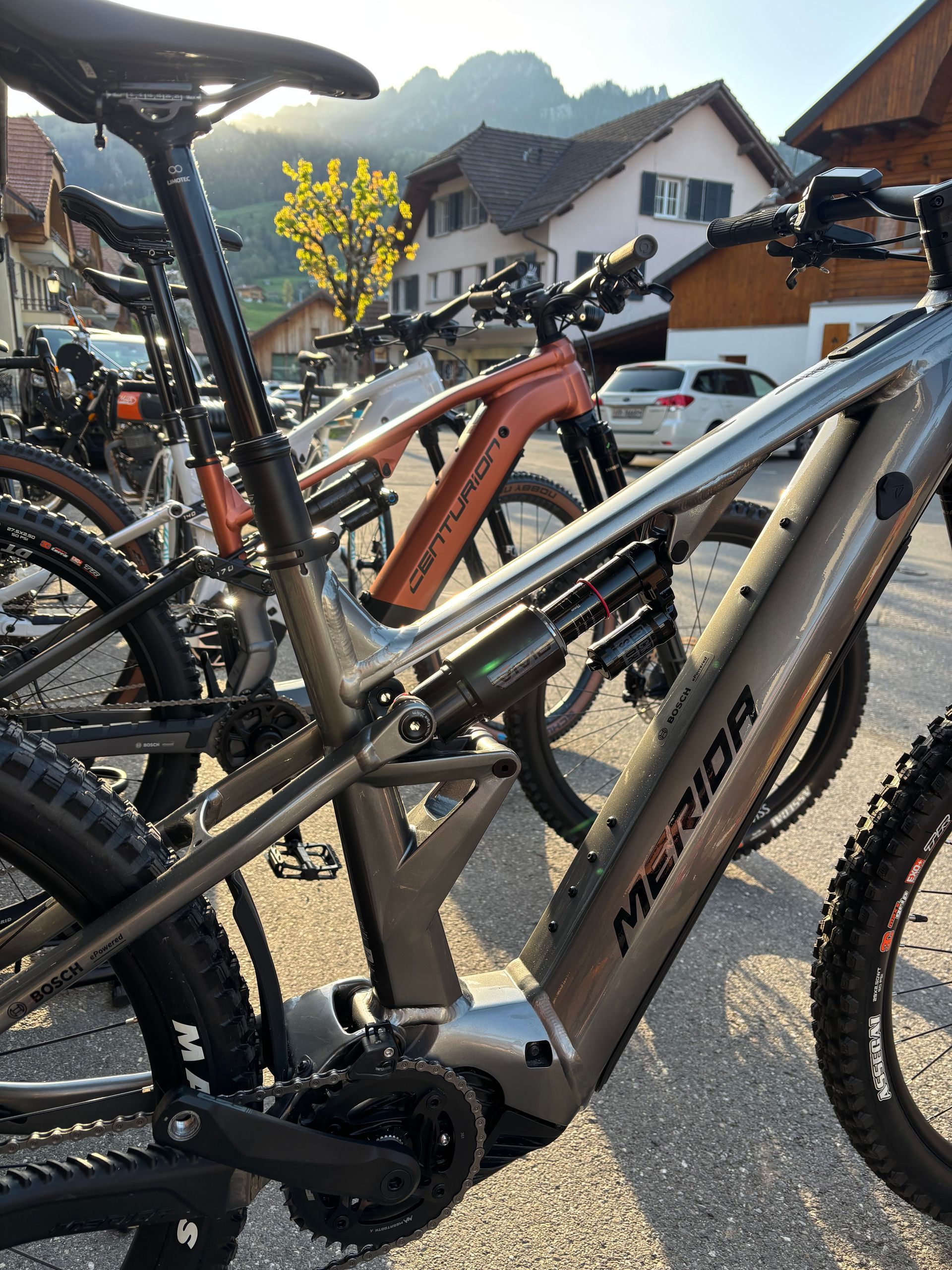 Vélo électrique Merida à cadre gris, garé à l'extérieur avec d'autres vélos, des bâtiments et des arbres en arrière-plan.