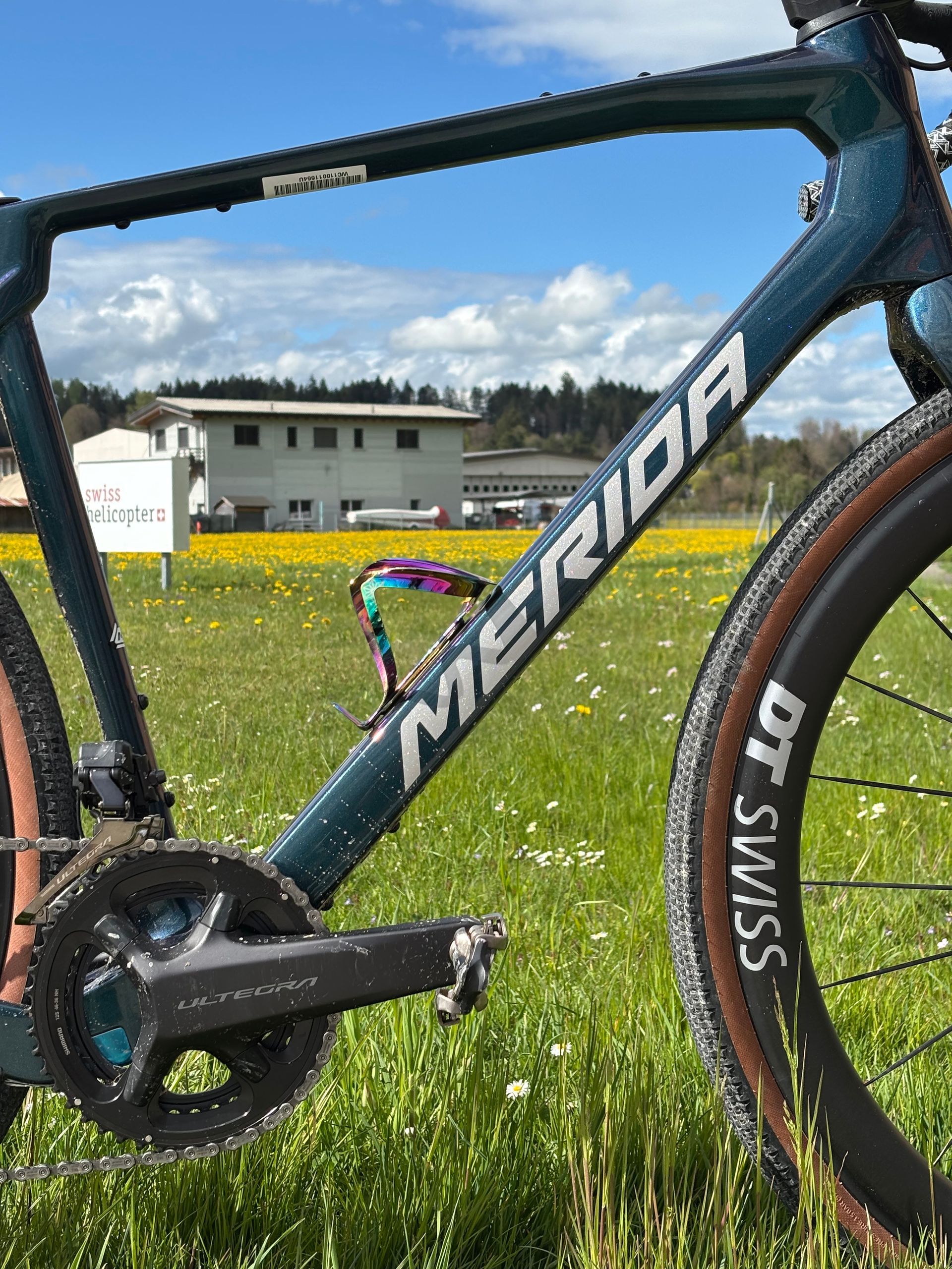 Vélo Merida turquoise sur herbe avec des fleurs jaunes, un bâtiment en arrière-plan. Pédalier noir et roue DT Swiss.