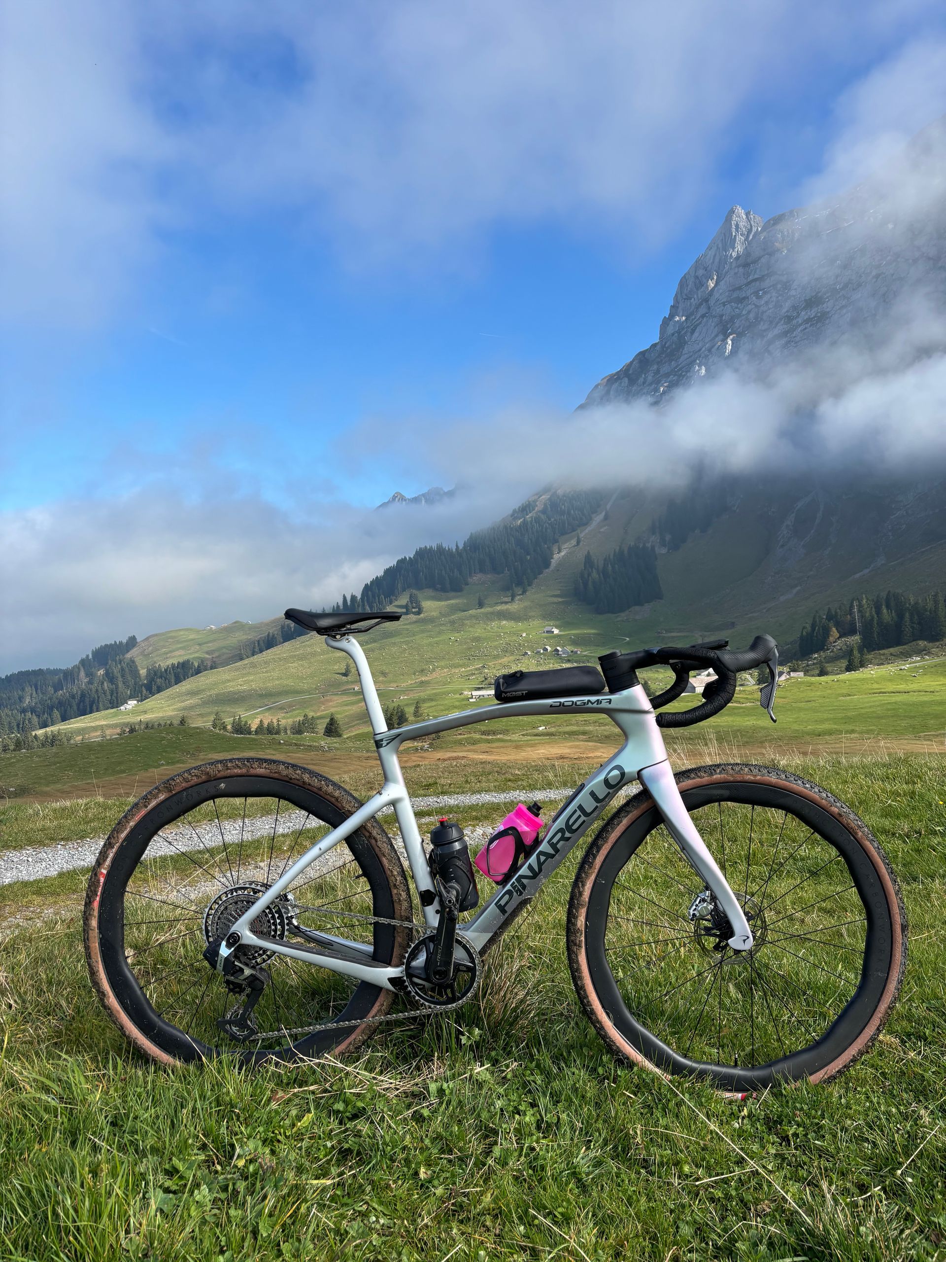 Vélo de gravel sur un coteau herbeux avec les montagnes en arrière-plan, sous un ciel nuageux.
