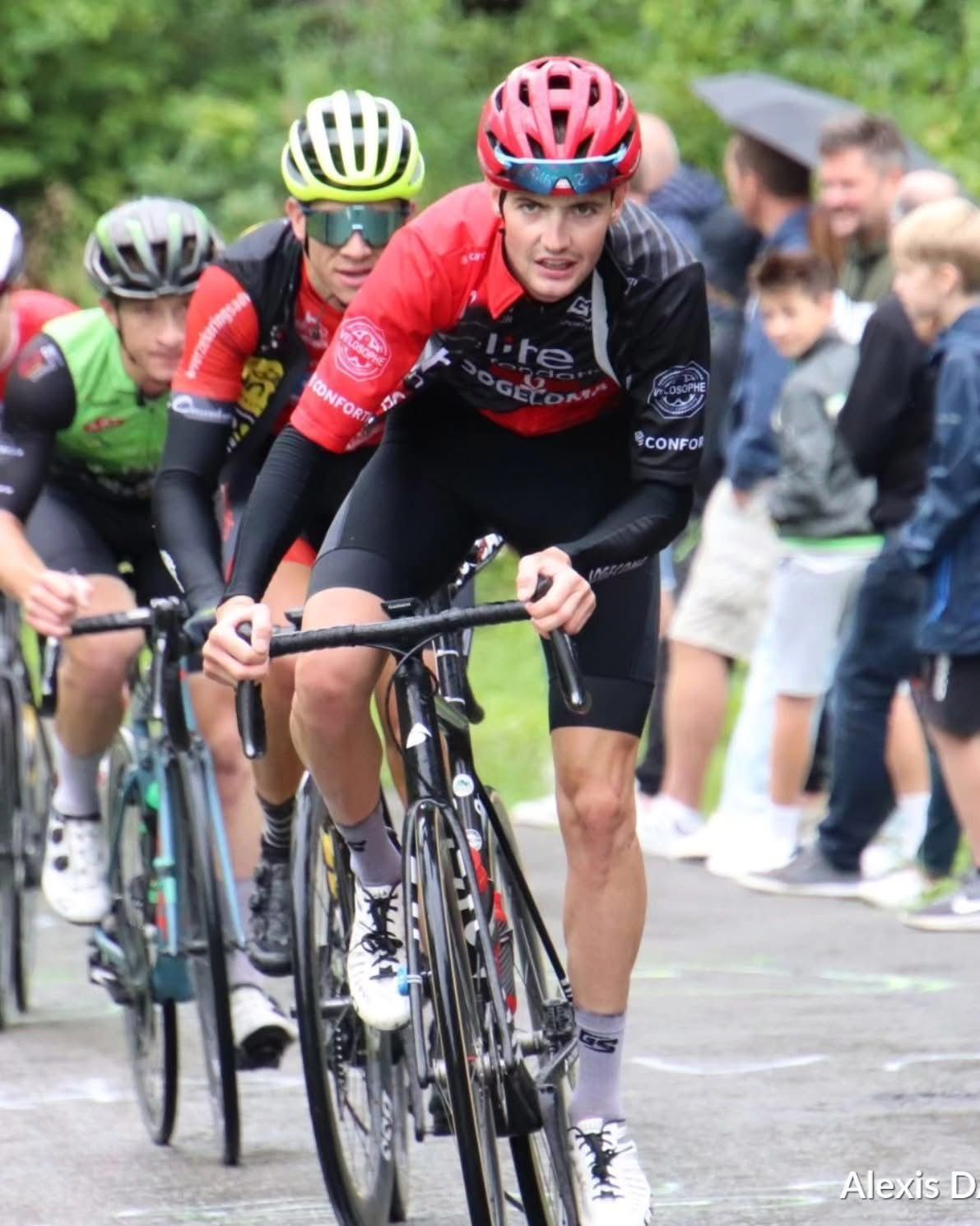 Cyclistes en course, coureur avec casque rouge, maillot noir et rouge, expression concentrée, sur une route, spectateurs en arrière-plan.