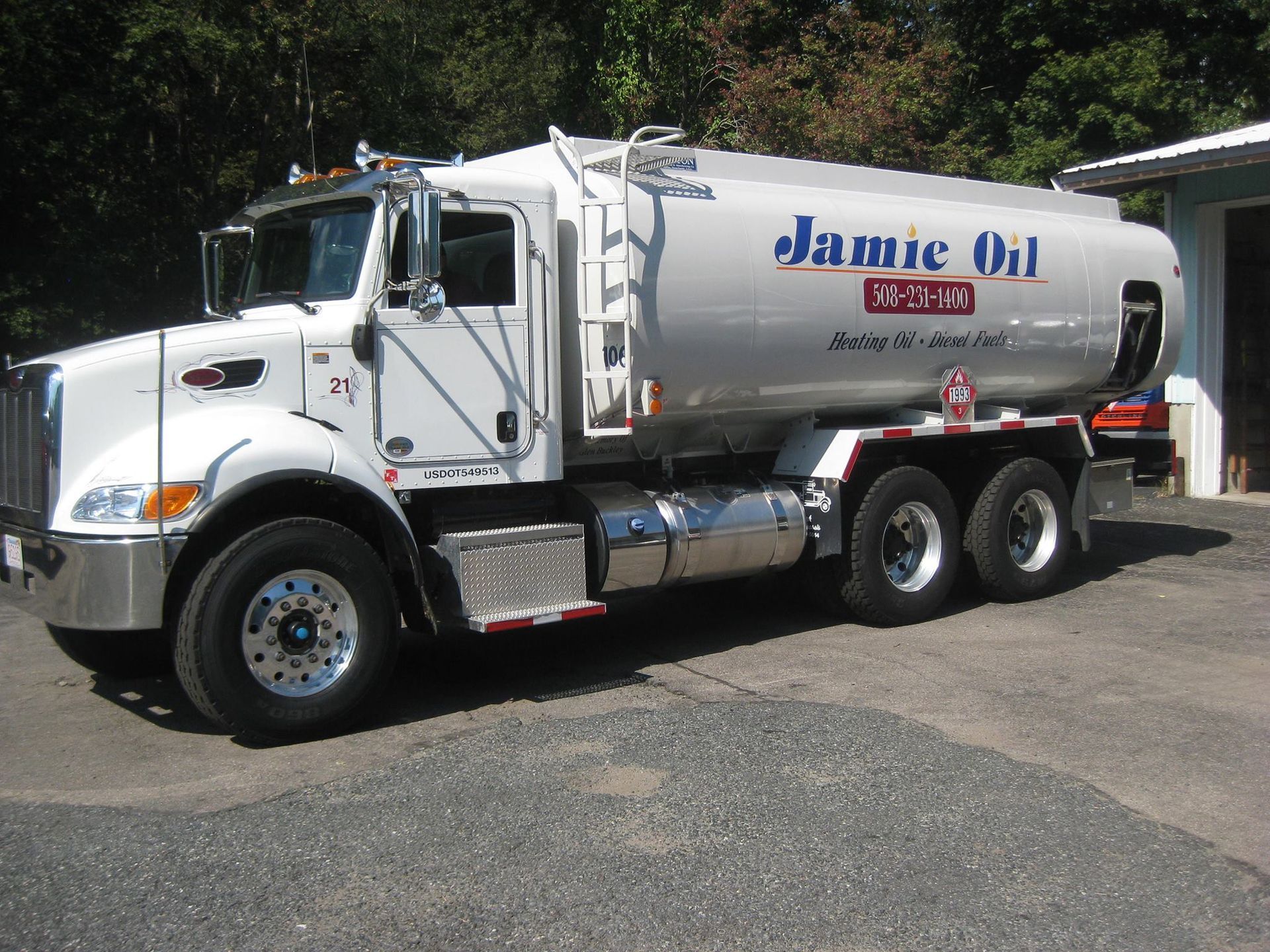 A white tanker truck drives on a multi-lane highway, sunny day, blue sky, trees in the background.