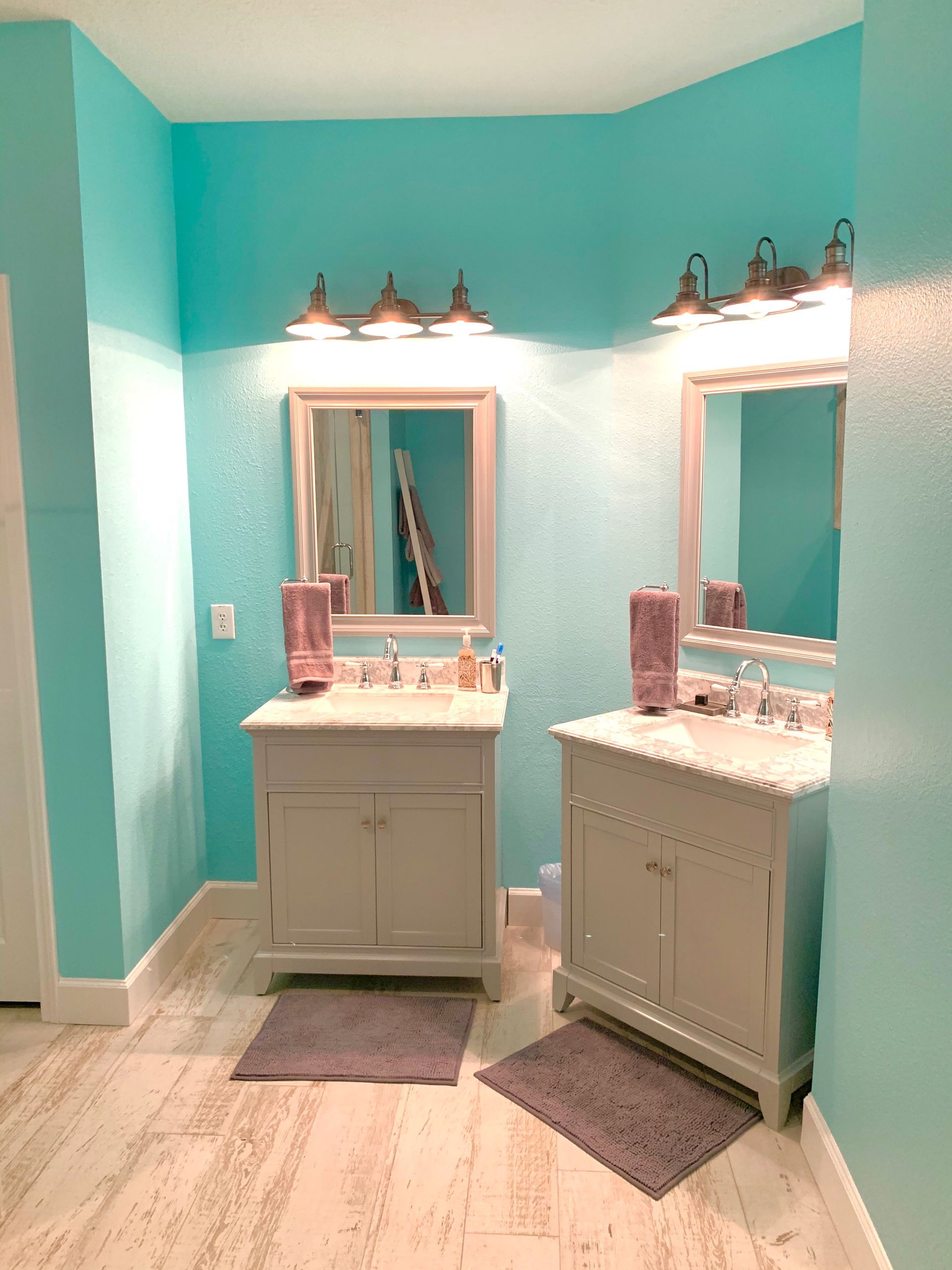 Two bathroom vanities with mirrors, under lights, in a room with light blue walls and light wooden floors.
