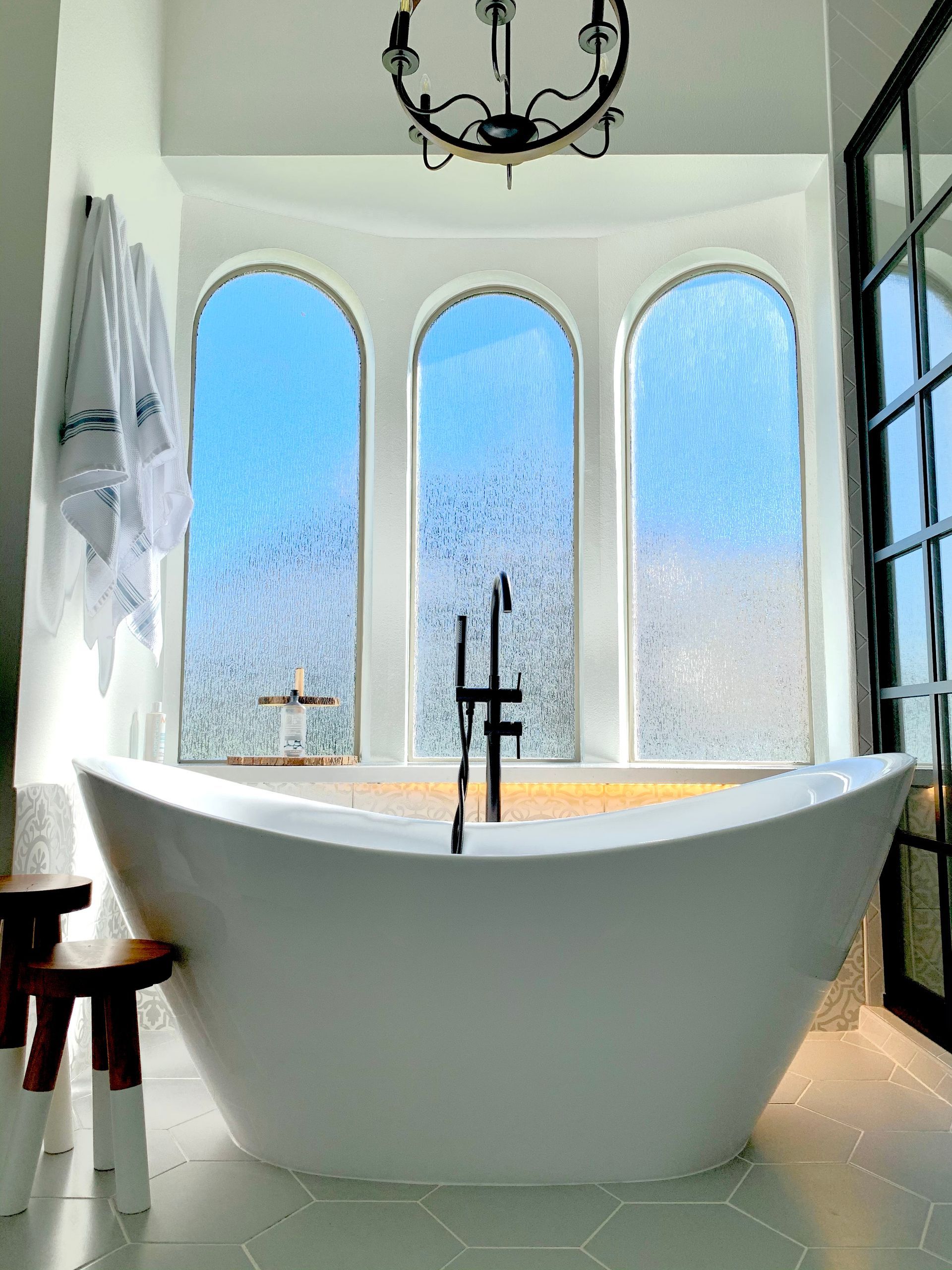 A white bathtub in front of three arched frosted windows with a blue sky view, a dark faucet, and a black chandelier.