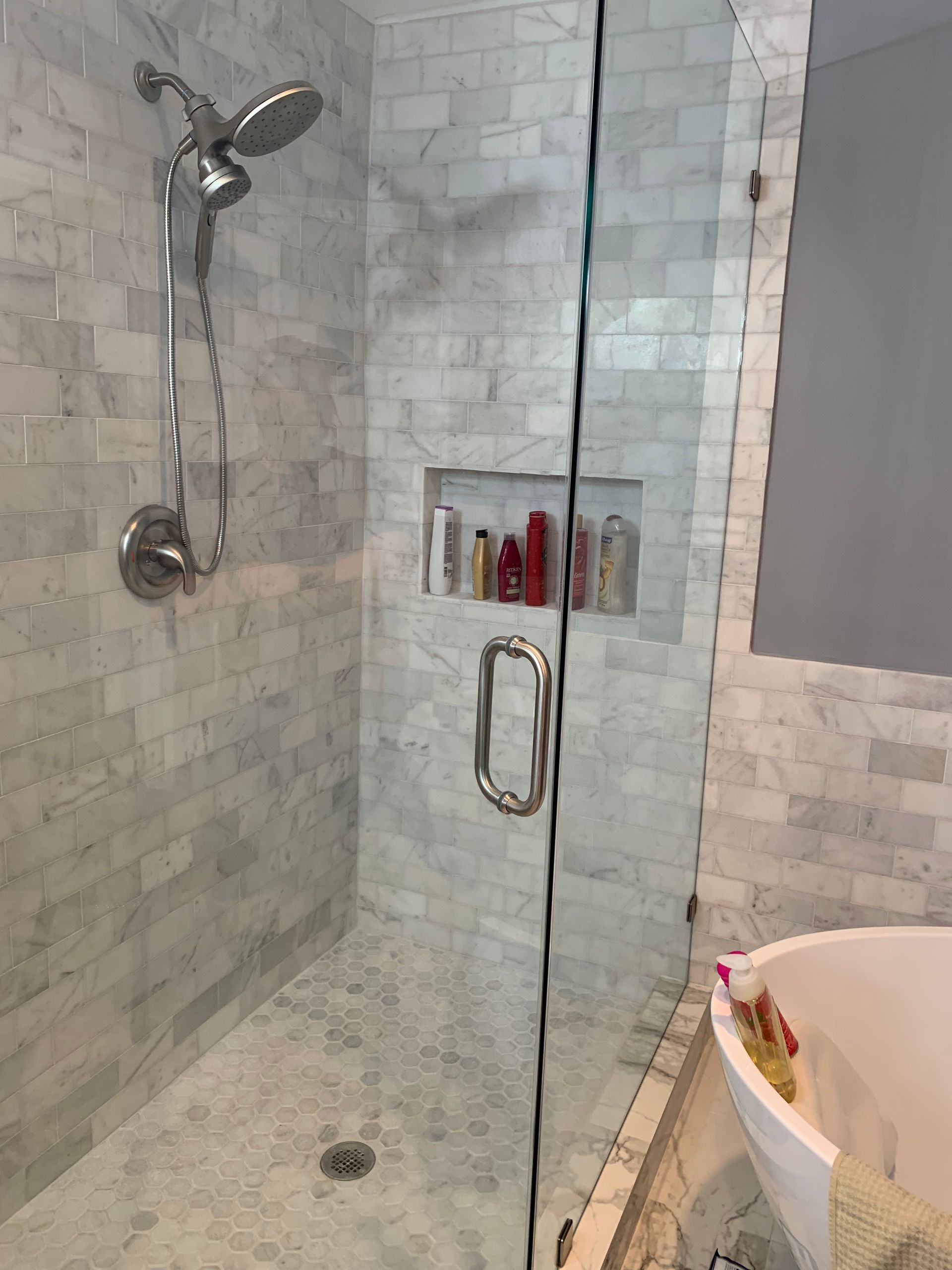 Shower with marble tile walls, glass door, and built-in shelf with shampoo bottles.