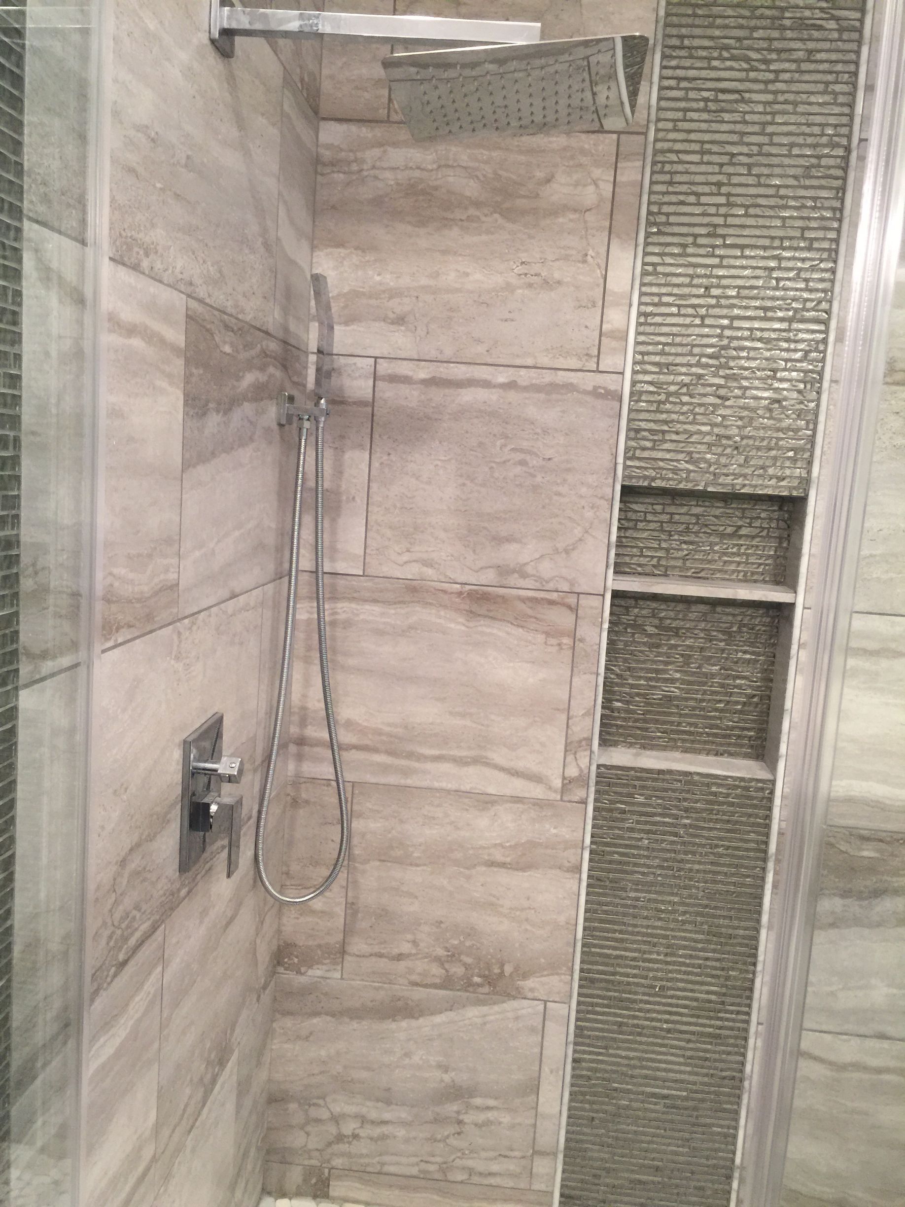 Shower interior with light gray, patterned tile, showerhead, handheld shower, and recessed shelf with textured tile.