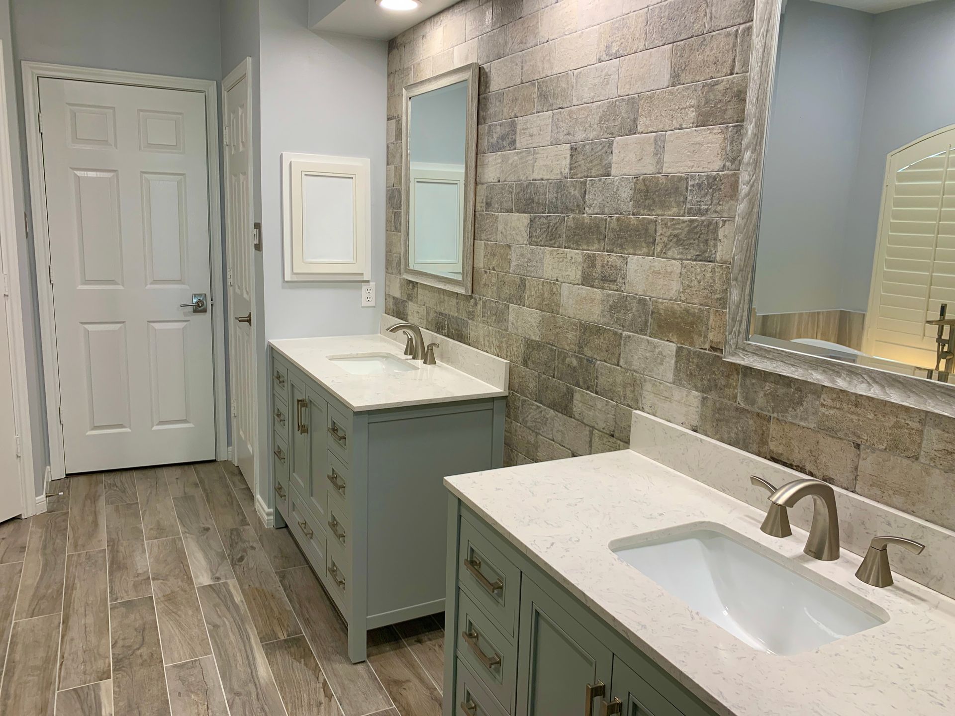 Bathroom with two sinks, gray cabinets, stone accent wall, large mirrors, and wood-look tile flooring.