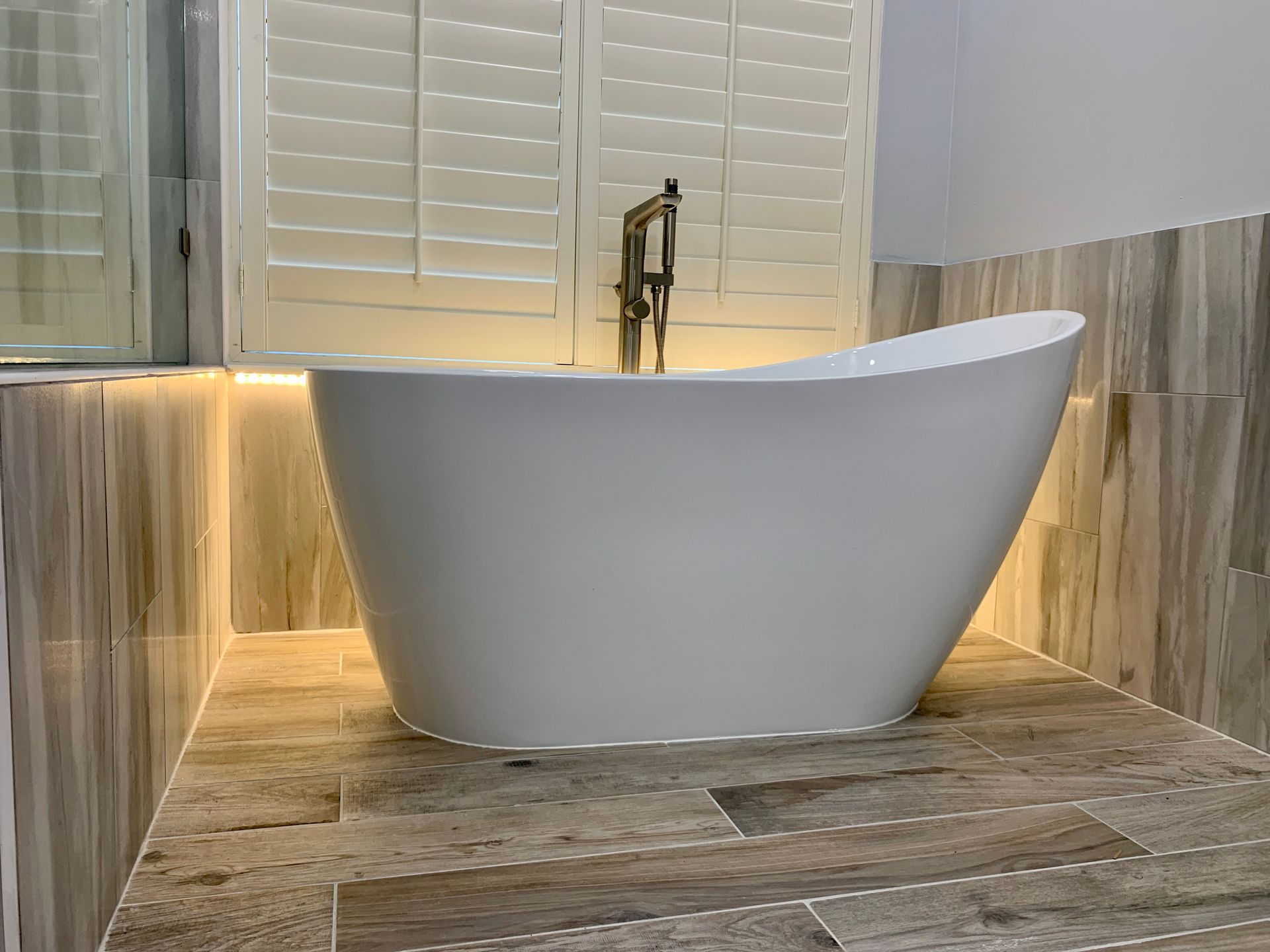 White oval freestanding bathtub with gold faucet in a modern bathroom with wood-look tile.