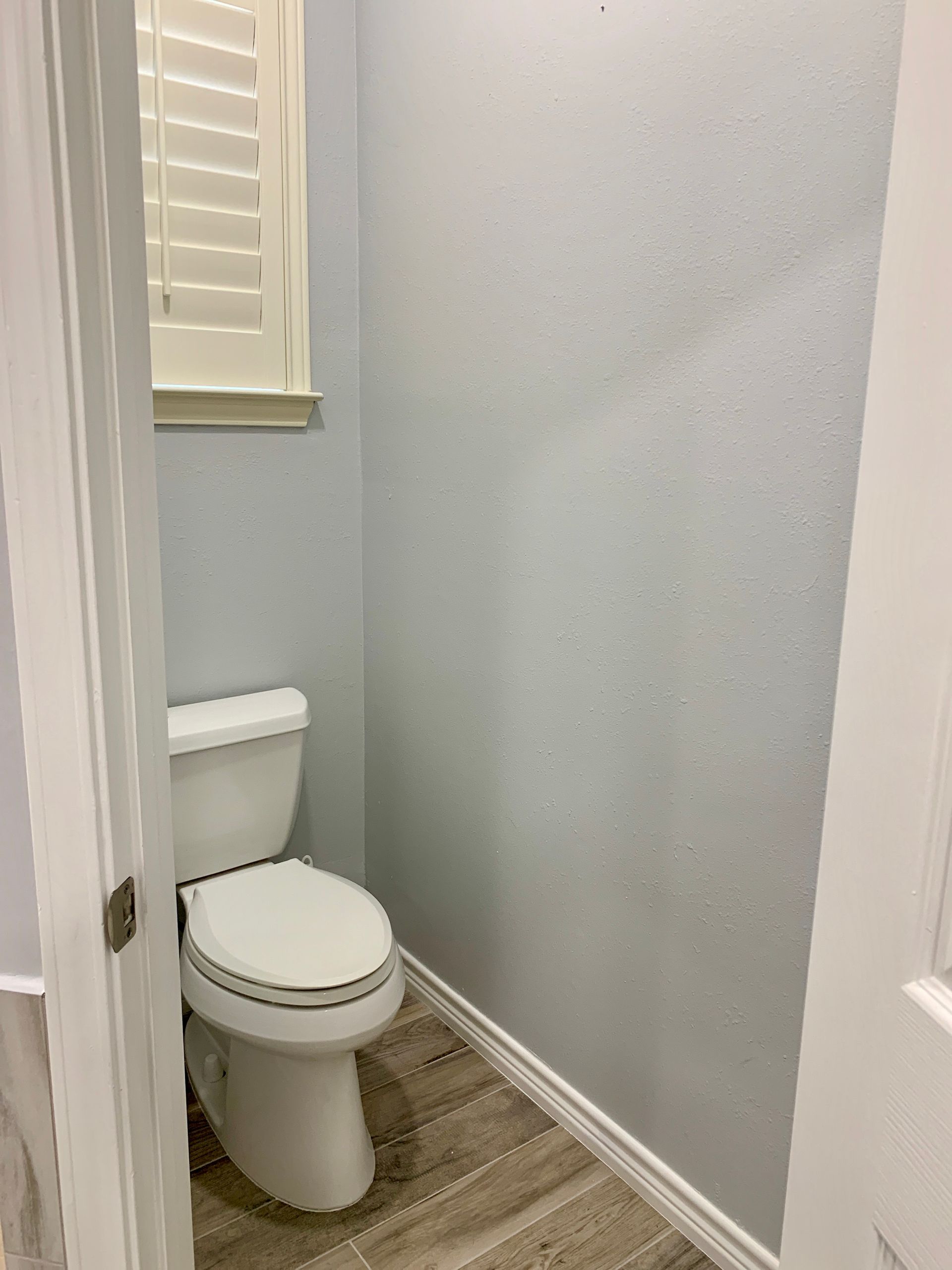 Toilet in a small bathroom with gray walls, a window with shutters, and wood-look flooring.