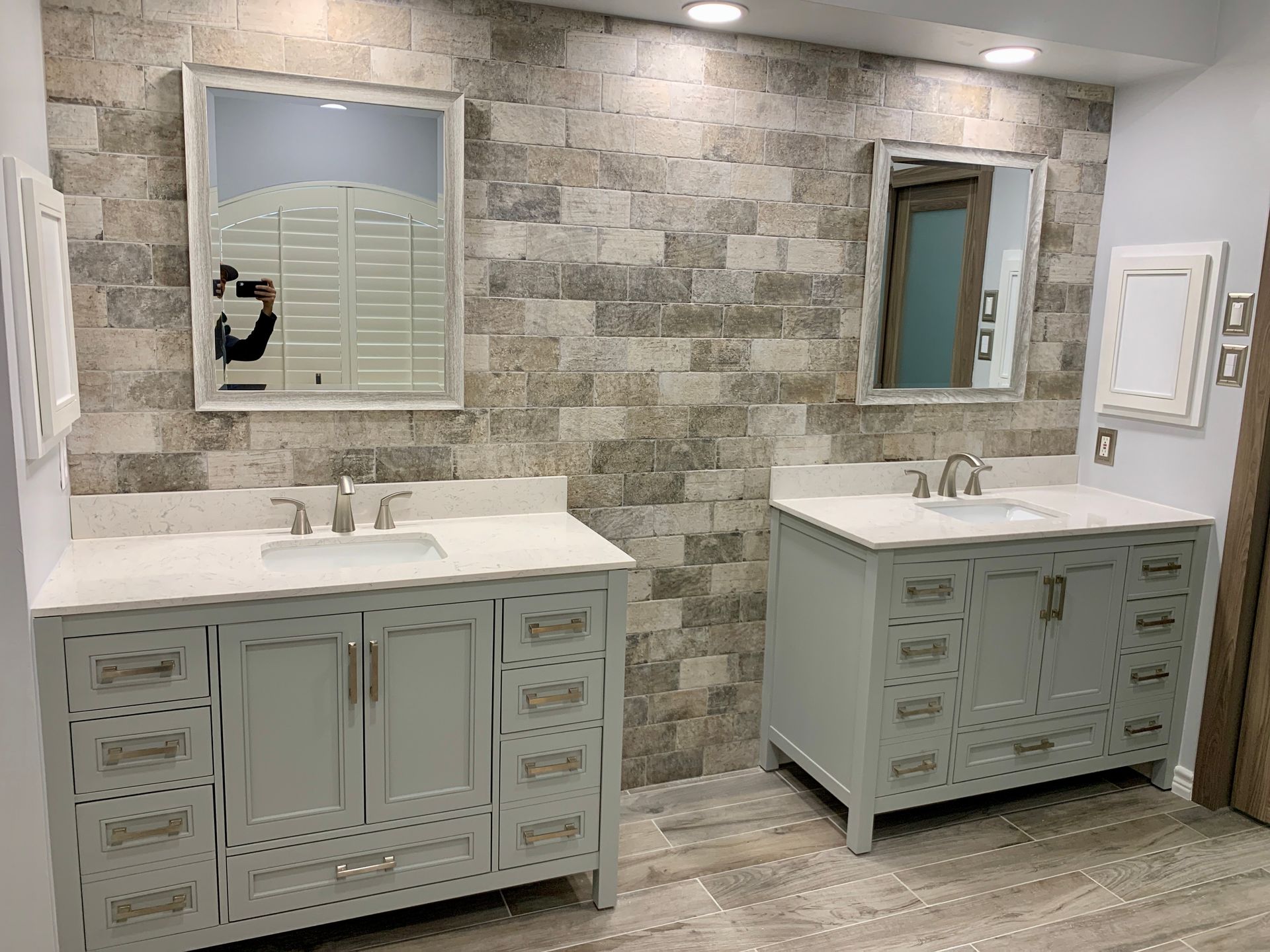 Two gray vanities with mirrors and sinks against a brick-like wall.