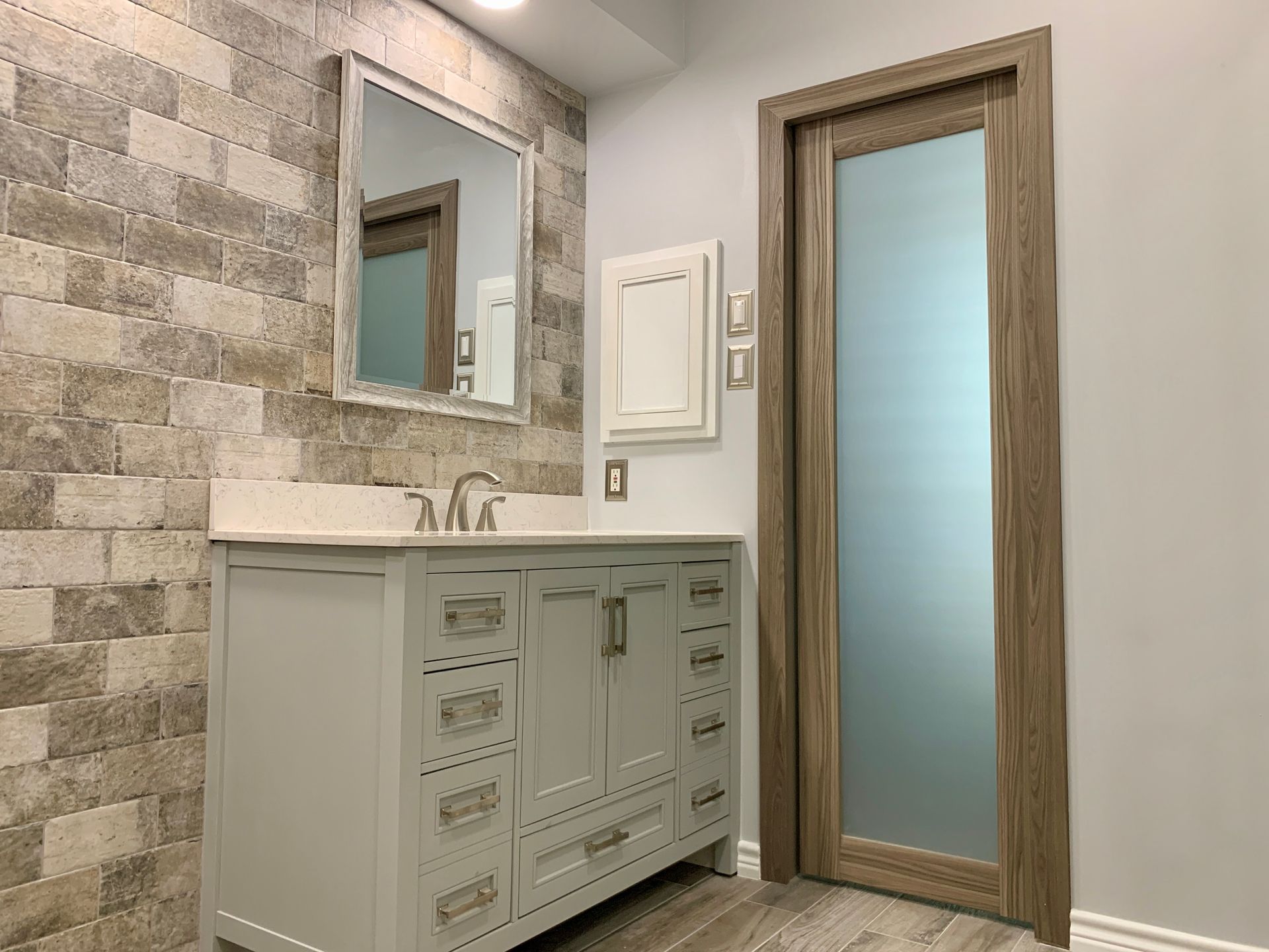 Bathroom with stone wall, light gray vanity, framed mirror, frosted glass door.