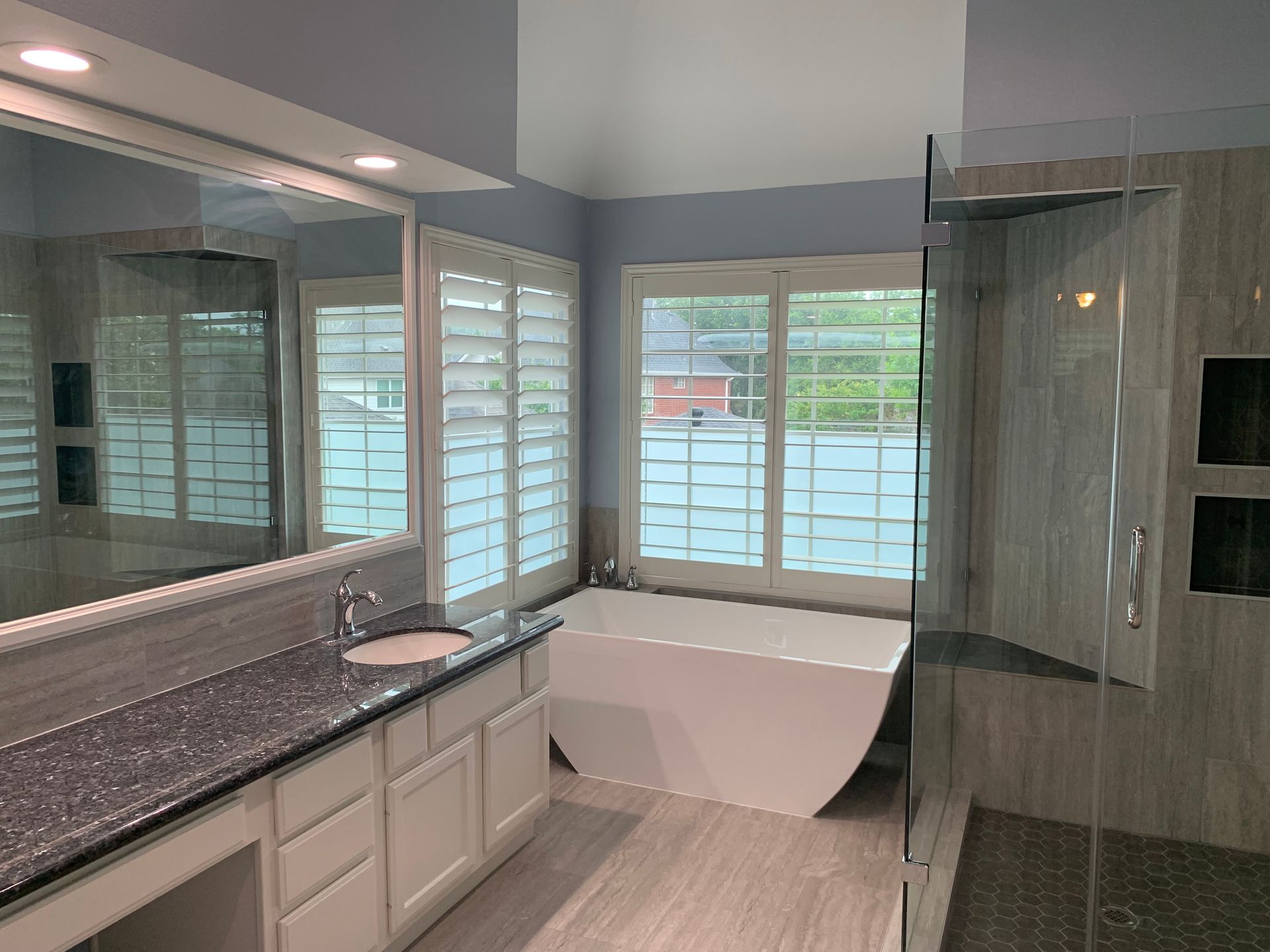 Bathroom with a white tub, granite countertop, glass shower, window shutters, and large mirror.