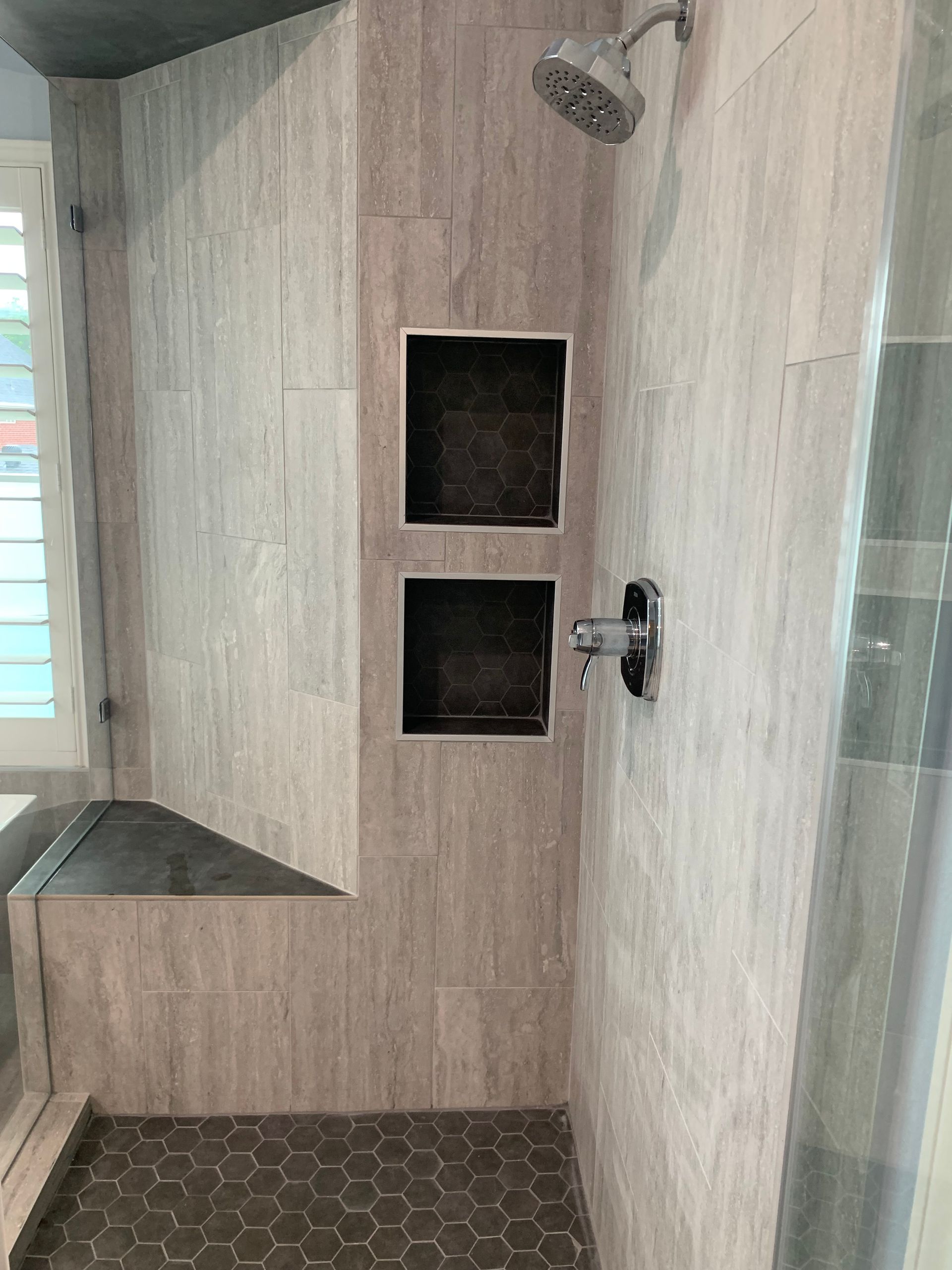 Shower with light gray tile walls, dark hexagon floor, built-in shelves, and a bench.