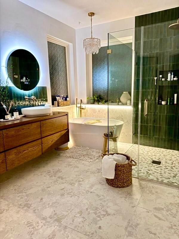 Luxurious bathroom with wood vanity, round mirror, soaking tub, and green tiled shower.