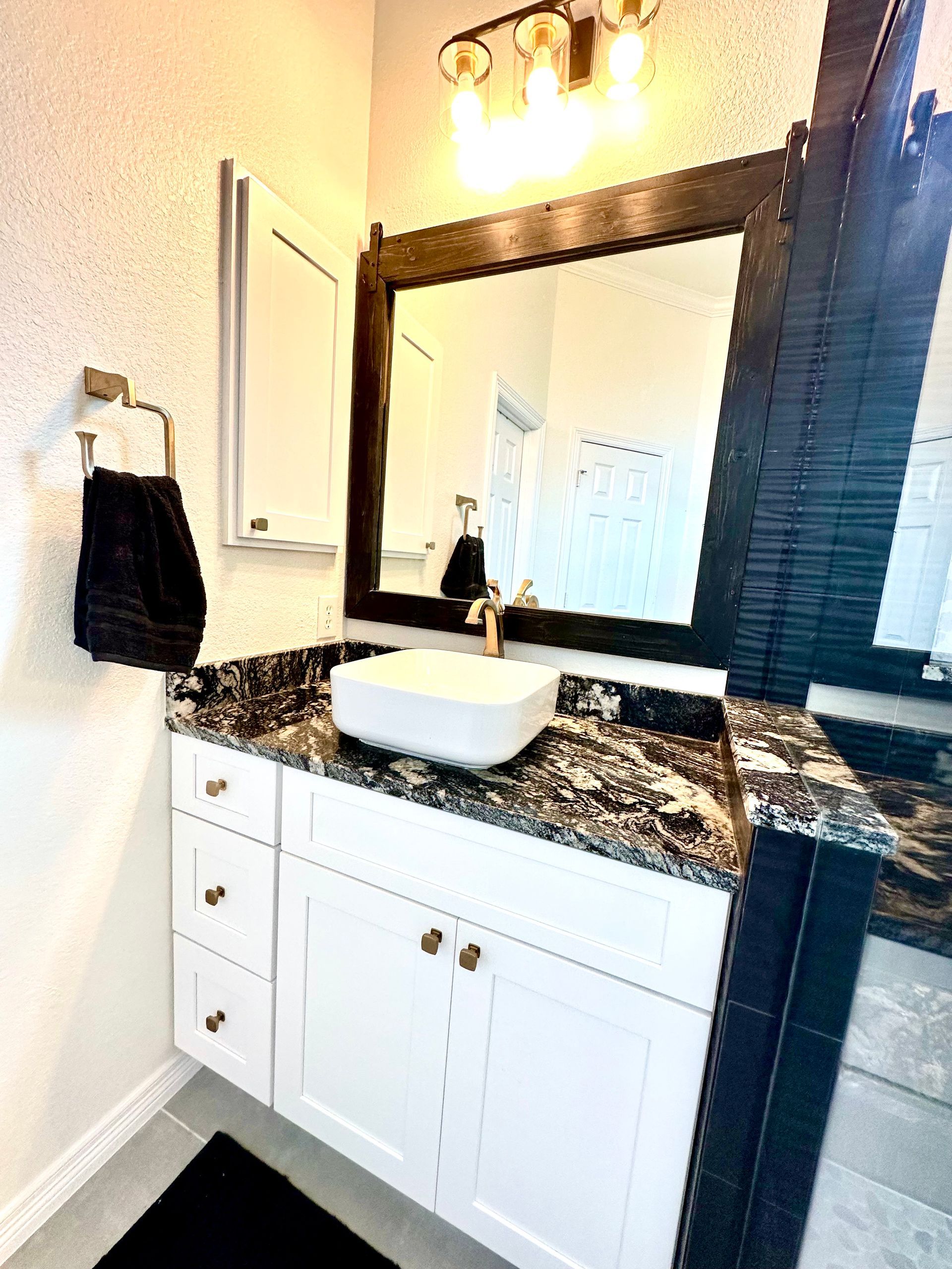 Bathroom vanity with white cabinet, black countertop, vessel sink, framed mirror, and gold fixtures.