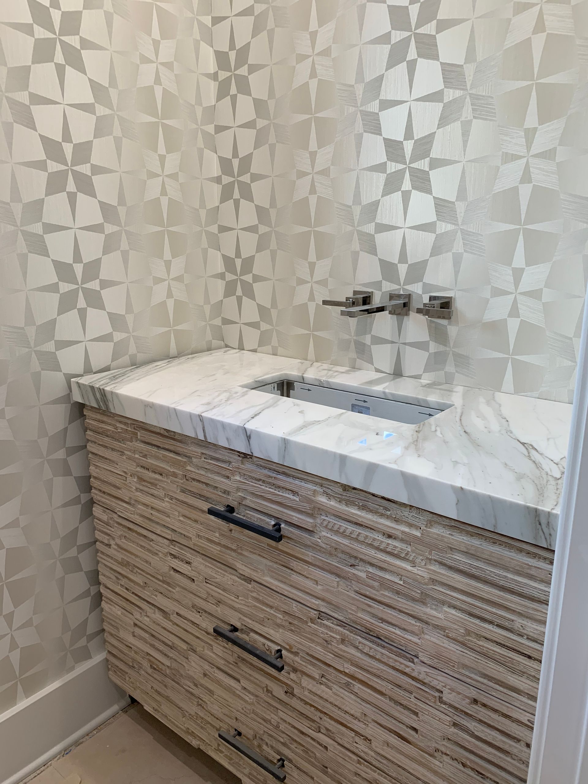 Light wood vanity with marble countertop and patterned wallpaper in a bathroom.
