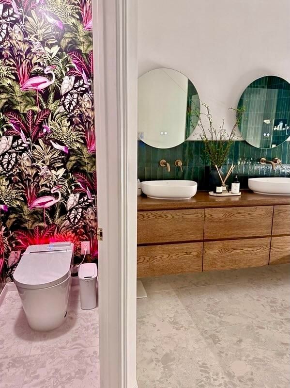 Bathroom with flamingo wallpaper, wooden vanity, and green tile accent wall. Two round mirrors.
