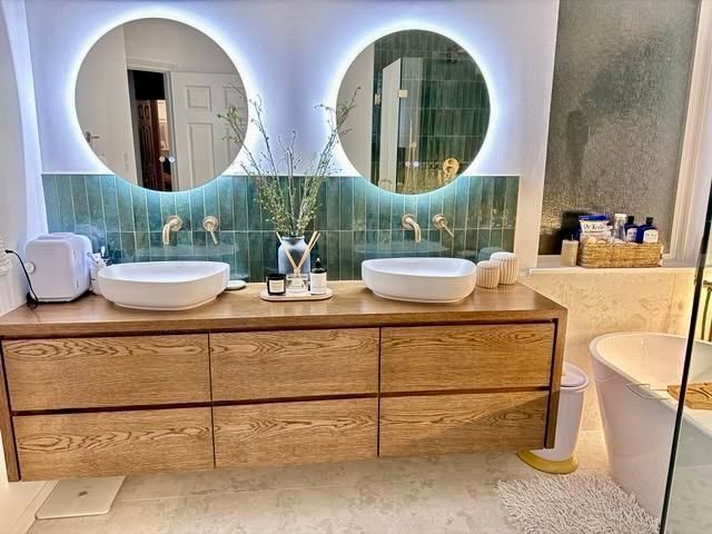 Modern bathroom with wooden vanity, two round mirrors with backlighting, and teal backsplash.