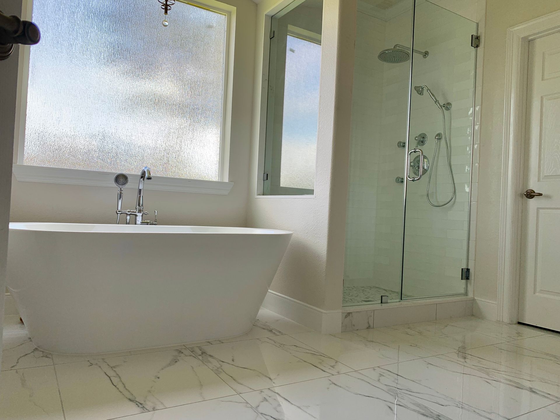 Bathroom with white freestanding tub, glass shower, marble tile, and textured window.