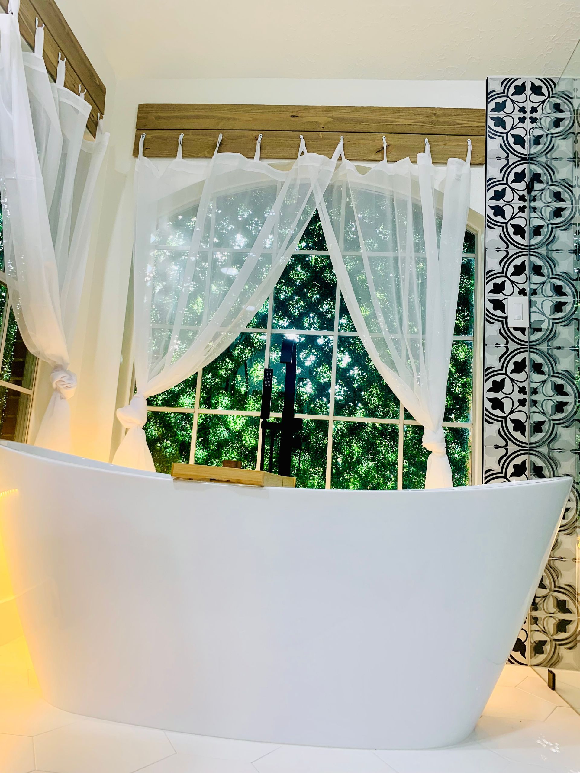 White bathtub in front of a window with white curtains, patterned tile wall.
