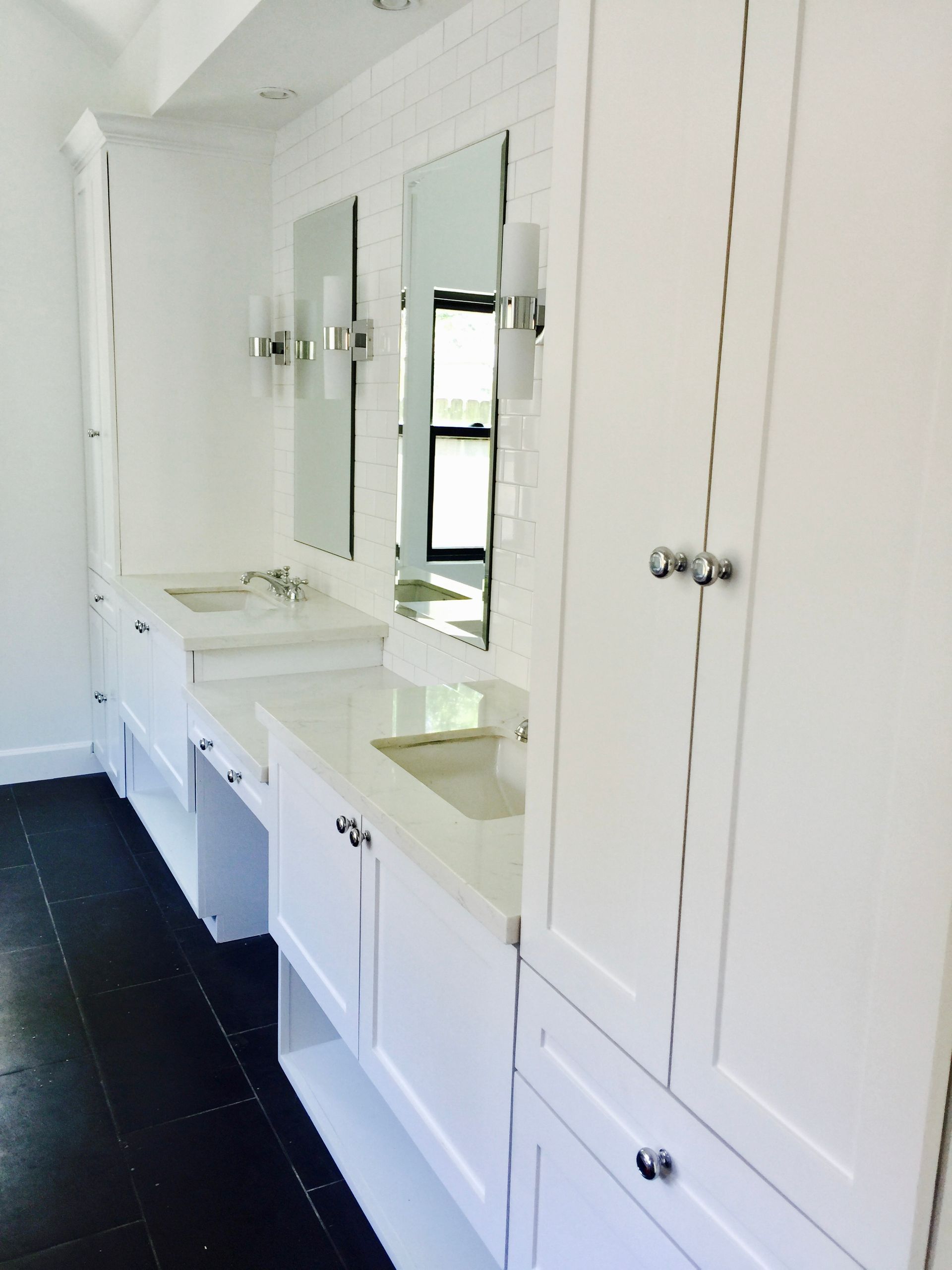 White bathroom with cabinets, mirrors, and sinks. Black floor, white brick wall.