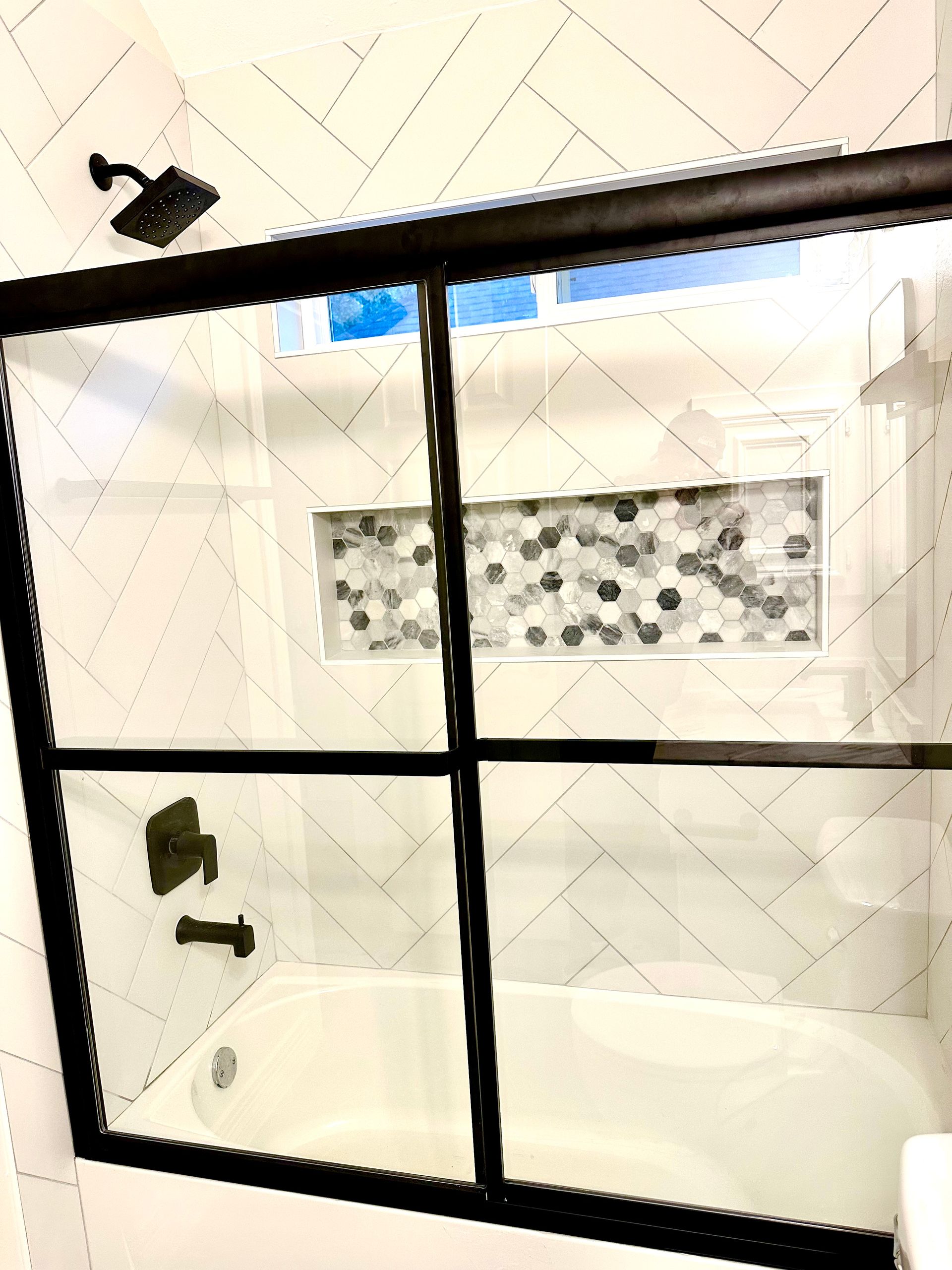 Bathroom with white herringbone tiles, black framed shower door, and bronze fixtures.