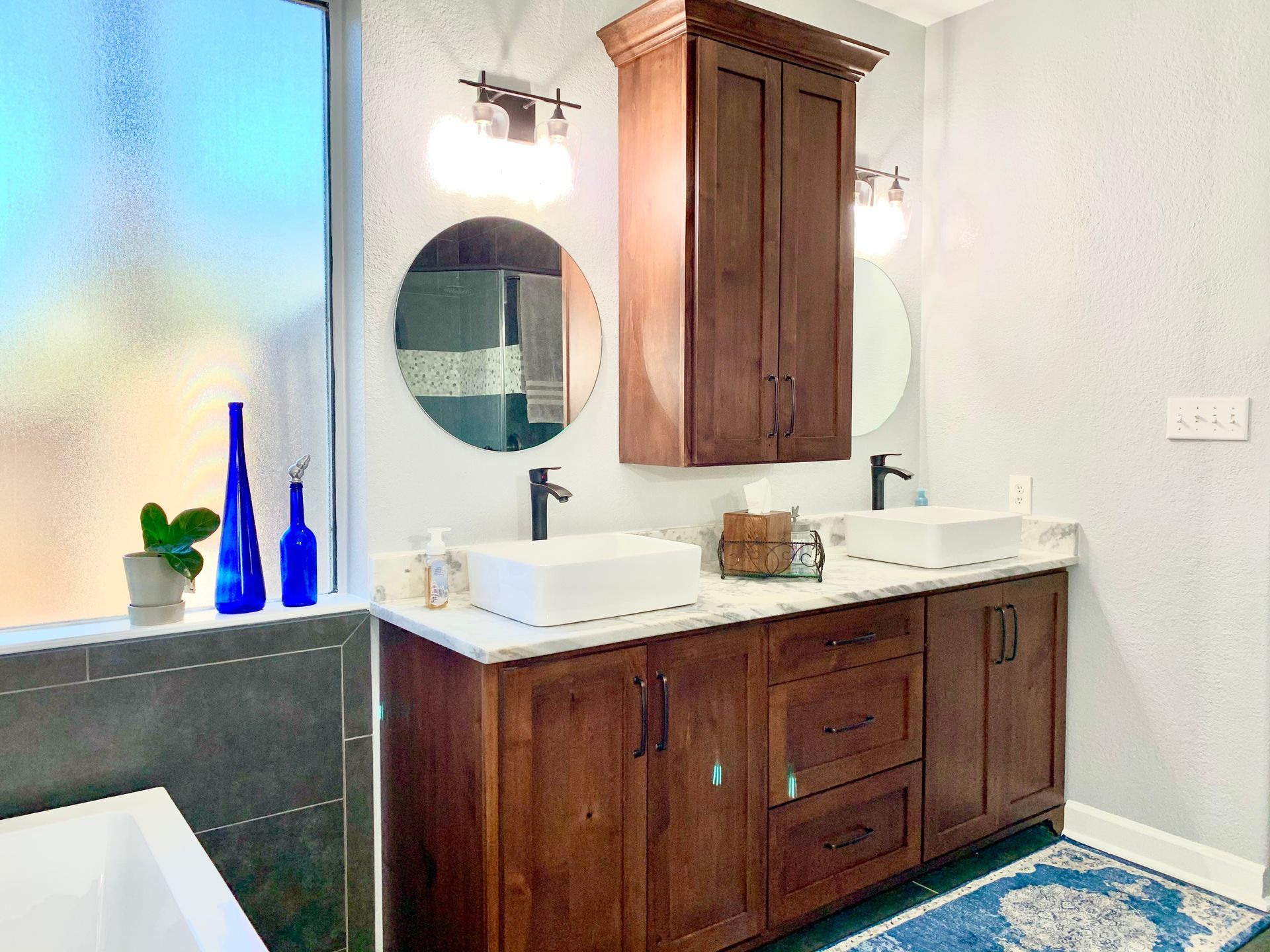 Bathroom with wooden vanity, two white sinks, round mirrors, and a frosted window.