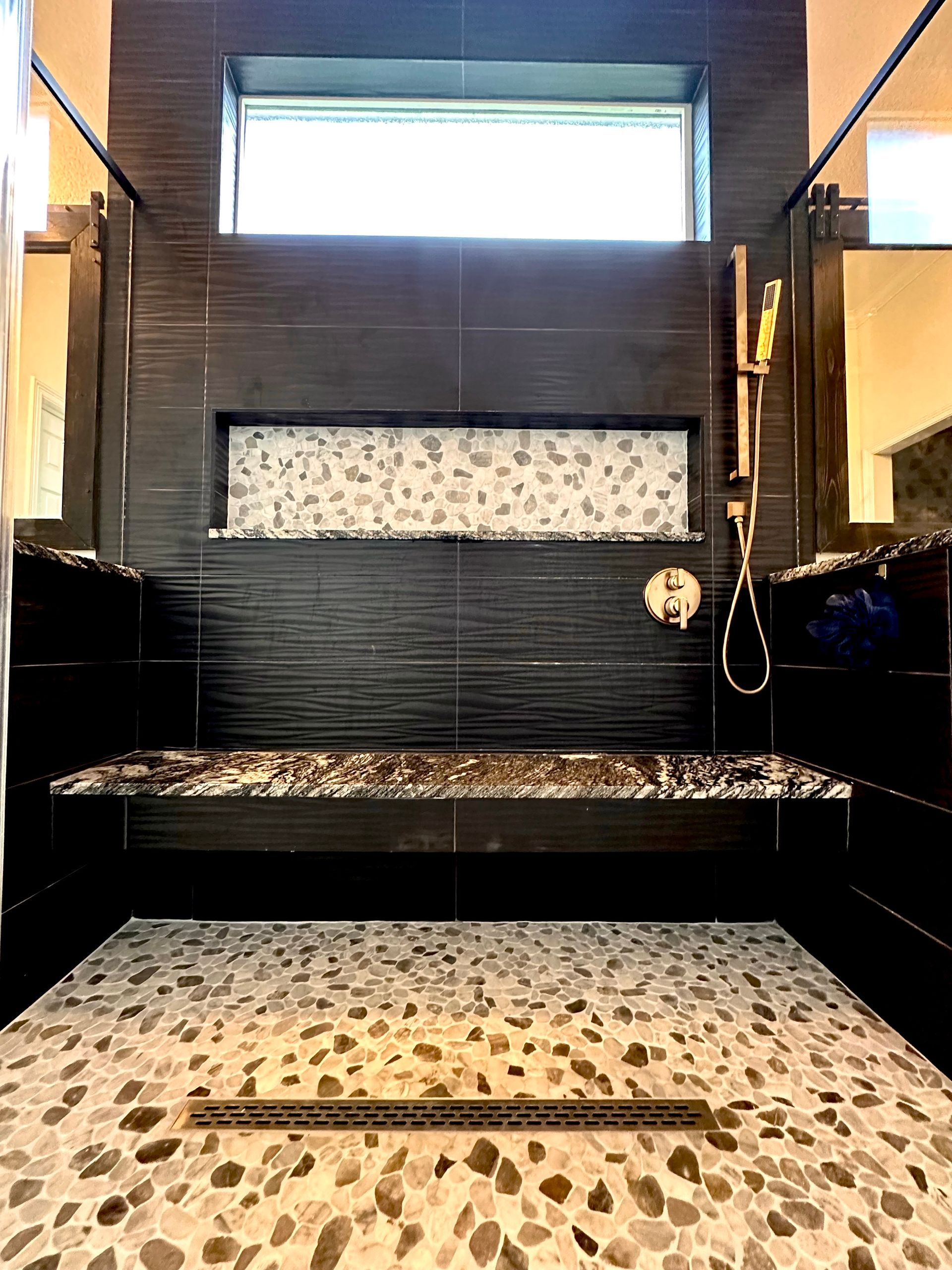 Shower with black tile walls, pebble floor, built-in bench, gold showerhead, and window above.