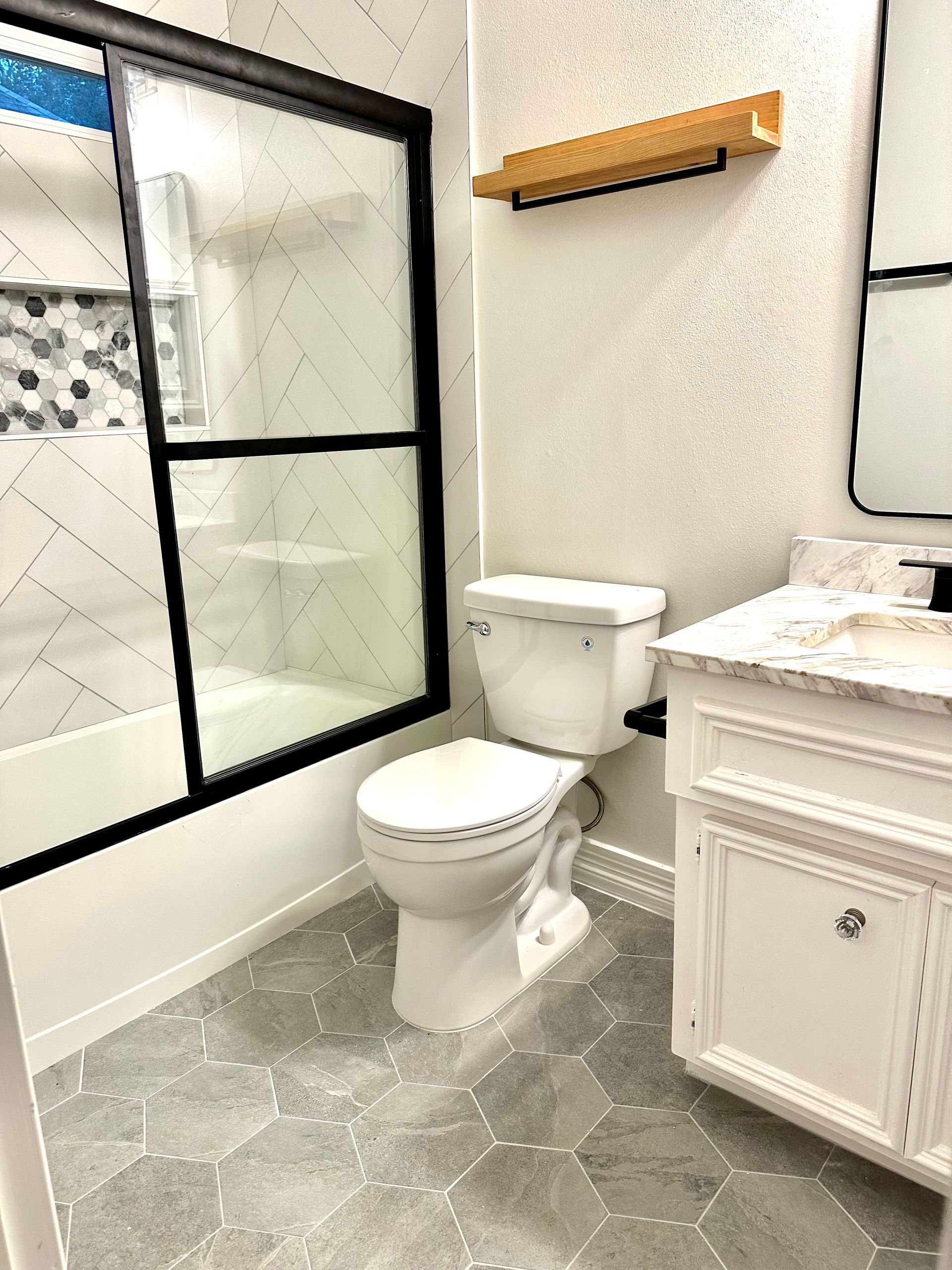Bathroom with white toilet, vanity, black-framed shower, and gray patterned floor.