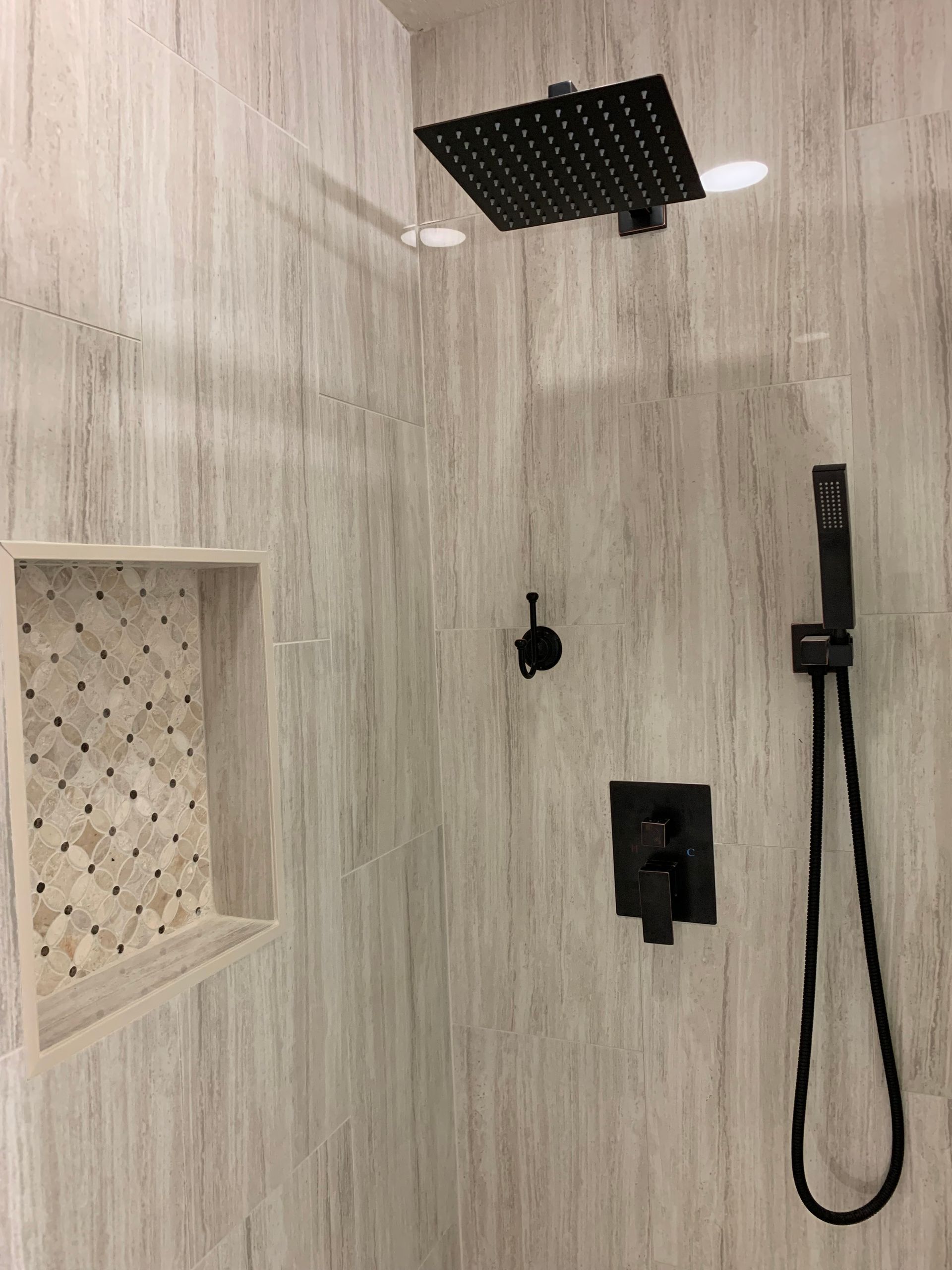 Shower with square black showerhead, handheld sprayer, and built-in shelf. Gray and white wood-look tile.