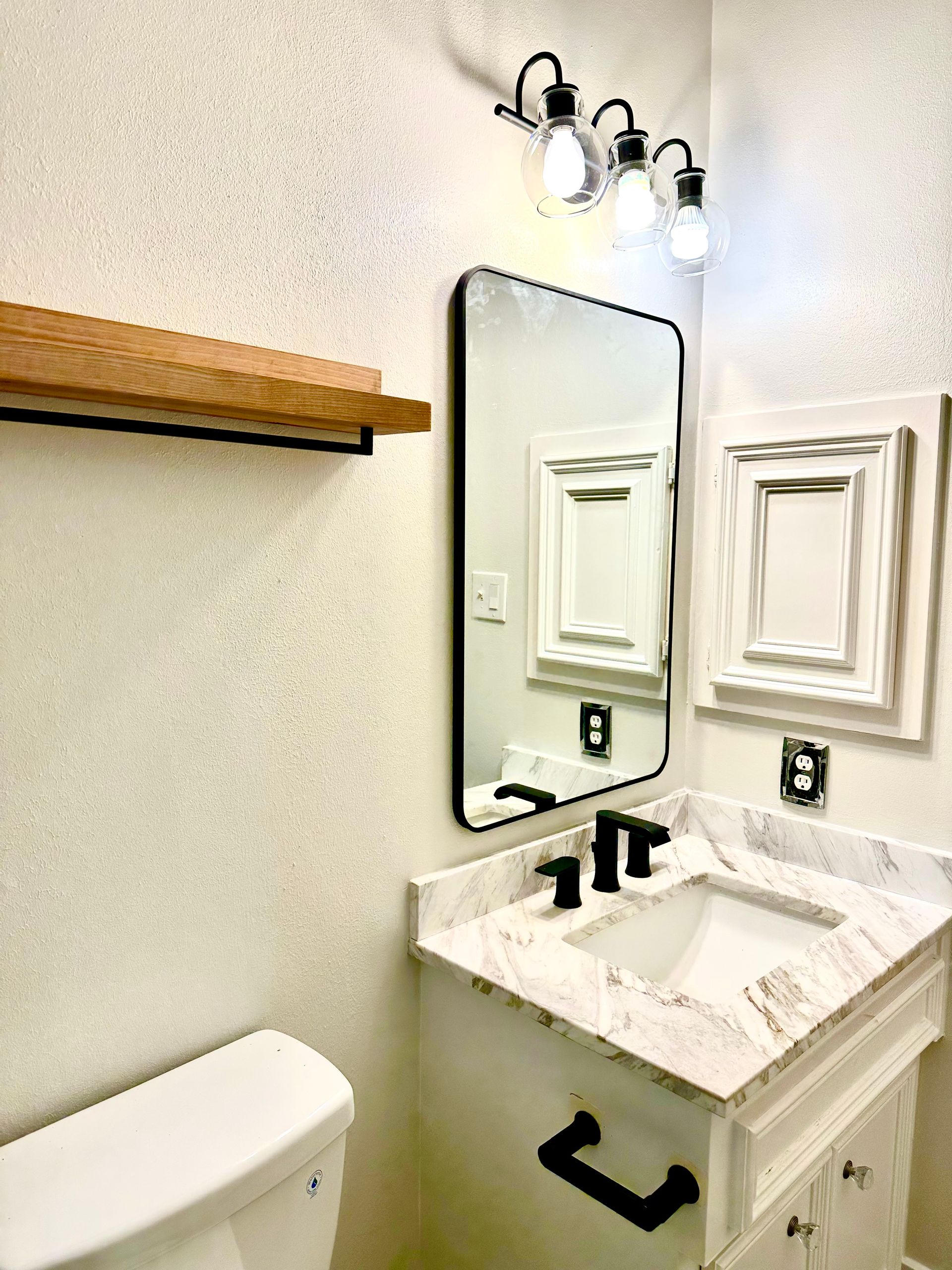 Bathroom with vanity, mirror, toilet, and wooden shelf on white textured walls. Black fixtures and light.