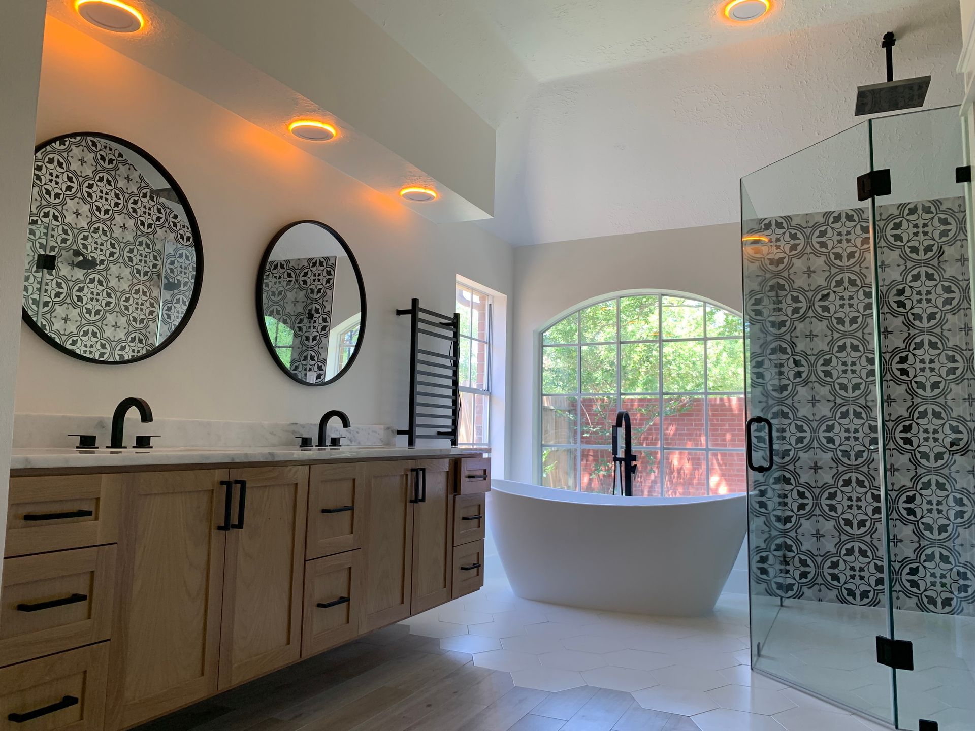 Modern bathroom with wooden vanity, black fixtures, and white freestanding tub near large window.